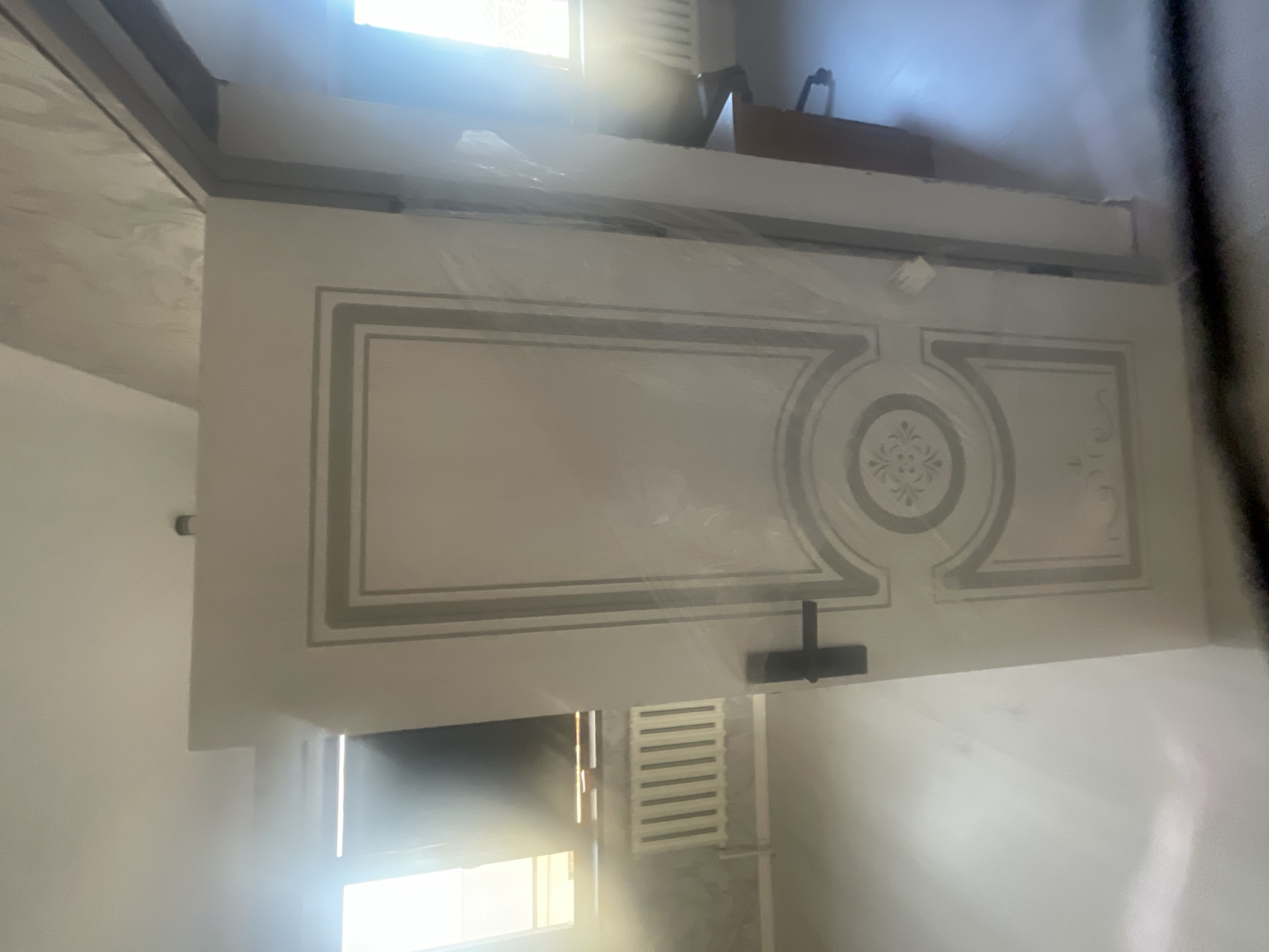 A slightly blurry, wide-angle, eye-level shot shows an interior scene with a closed white door with a black handle on the left. The door has decorative molding. To the left of the door, a window with light coming through is visible, and below it is a white radiator. The floor is a light color, and to the right of the door, the scene opens into another room. Part of a desk and an office chair are visible in the background on the right. The overall impression is of a room that is perhaps under renovation or has recently been furnished. The location is not discernible from the image itself.