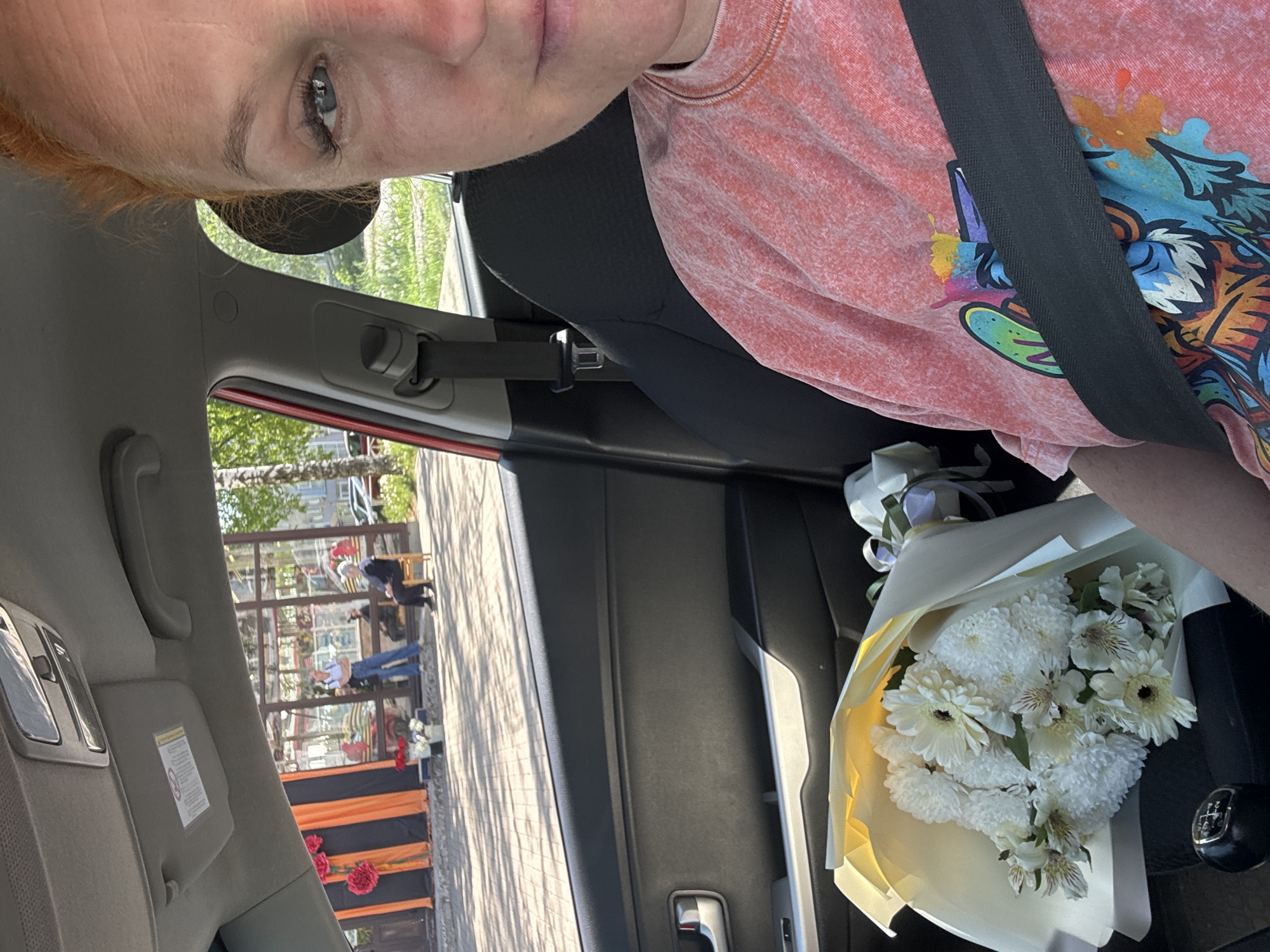 A selfie taken from the driver's seat of a car shows a woman wearing a seatbelt and a pink t-shirt with a cartoon design. In the passenger seat is a bouquet of white flowers wrapped in yellow paper. Through the open car window, a street scene is visible with a building decorated with orange and black striped banners and red flowers. Two people are standing outside the building, one seated. Based on the decorations and presence of people in formal attire, the scene suggests a public memorial event or celebration in Chaikovsky, Russia.