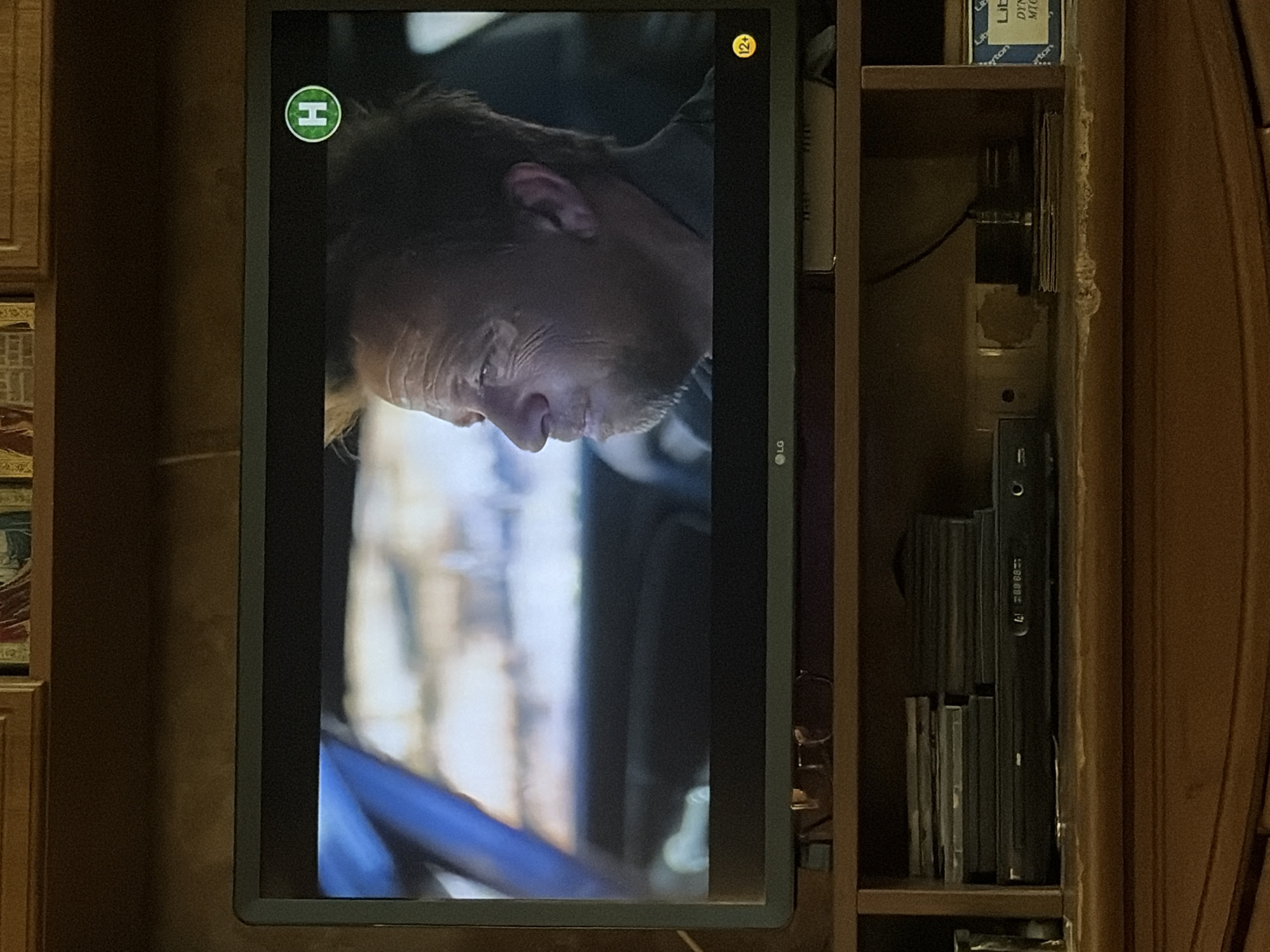 A television screen displays a man's face in profile. The man is light-skinned with short, blondish-brown hair and appears to be middle-aged. He is wearing a dark collared shirt. A green circle with a white "H" is visible in the upper right corner of the screen. A yellow circle with "12+" is visible in the lower right corner of the screen. The LG logo is visible on the bottom bezel of the television. Below the television, a DVD player is visible. The television and DVD player are on a wooden shelf, which is part of a larger wooden entertainment unit. The background is dark and appears to be a wall.