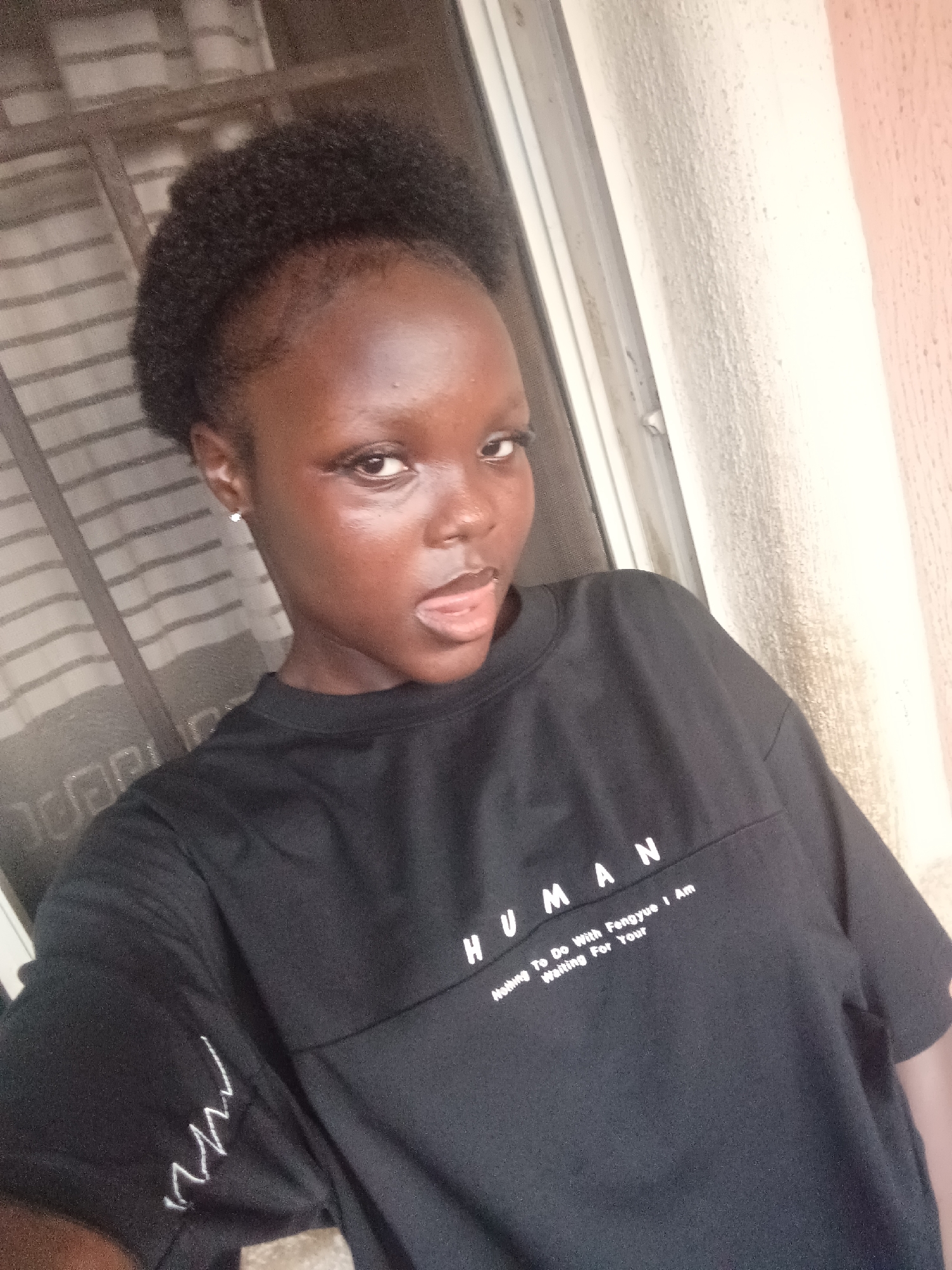 A young, dark-skinned woman is posing for a selfie. She is wearing a black T-shirt with the word "HUMAN" printed on it, along with smaller text that reads "Nothing To Do With Fengyue I Am Waiting For Your." The woman's hair is styled in a short, afro-like manner. She is wearing a small earring in her left ear. She has a serious expression, with her tongue slightly protruding.
The background is composed of a window with striped curtains and a textured wall. Based on the complexion of the woman, the location is estimated to be somewhere in Onitsha, Nigeria. There is no visible interaction or action beyond the subject posing. The surrounding environment appears to be an indoor space, possibly a home.