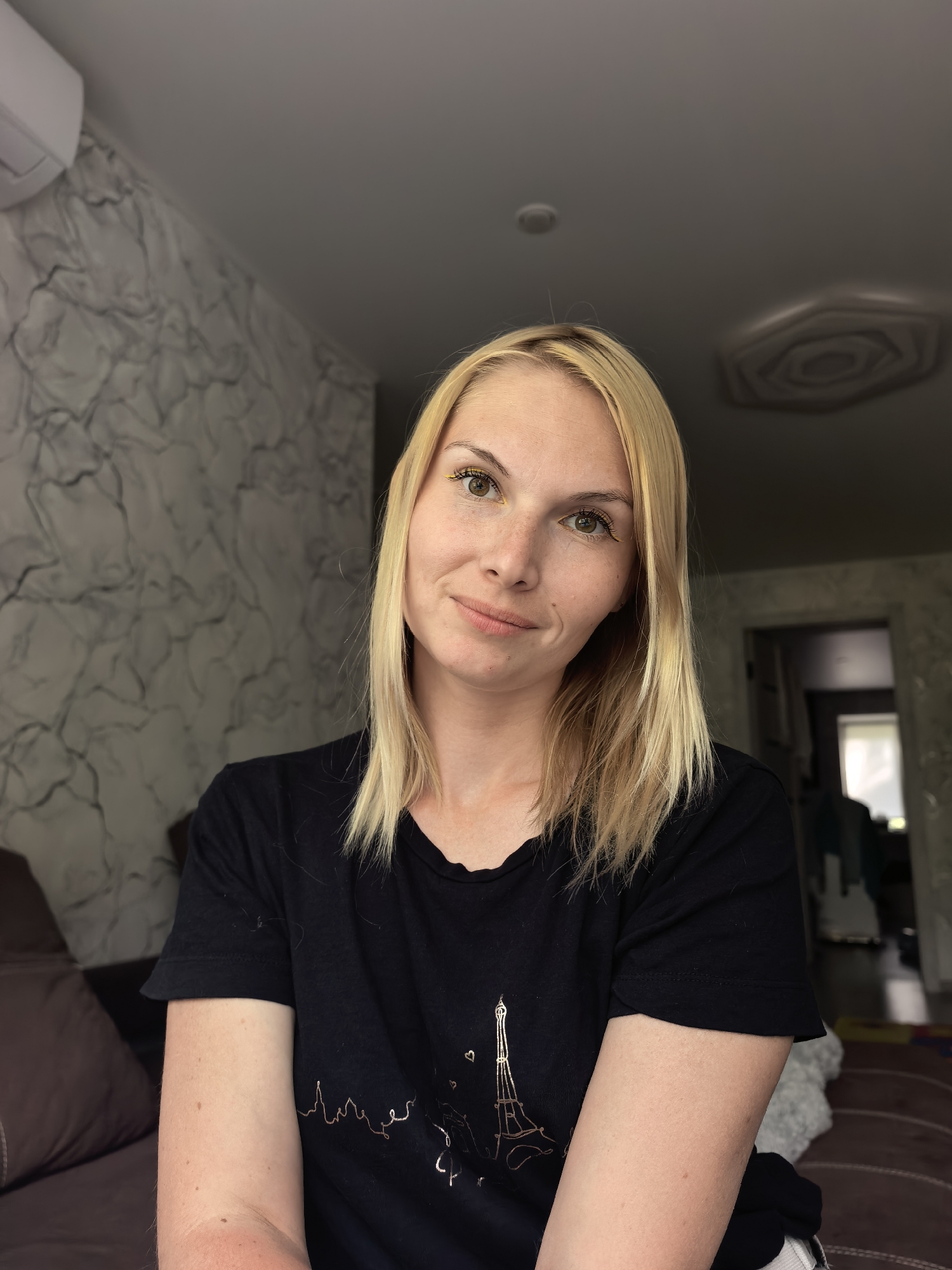 A medium shot image shows a fair-skinned woman with blonde hair and green eyes, looking directly at the camera. She is wearing a black t-shirt with a graphic of the Eiffel Tower. The woman has light makeup on, including yellow eyeliner. She has freckles across her nose and cheeks and a slight smile. The background appears to be an indoor living space with a textured grey wall, a brown couch, and a doorway leading to another room. The lighting suggests it is daytime. The image was taken in Sterlitamak, Russia.