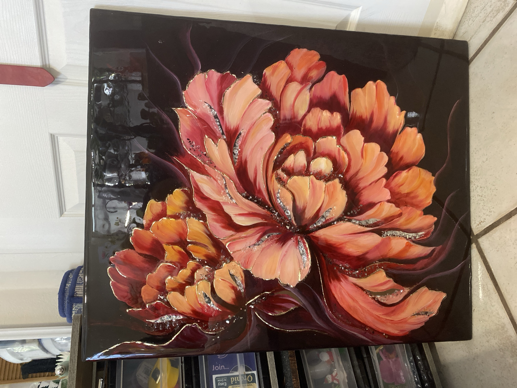 The image shows a close-up of a painting featuring two large, abstract flowers in shades of red, orange, and pink. The petals are richly textured and highlighted with silver glitter and gold trim. The background is a dark, glossy black, creating a dramatic contrast. The painting is mounted on a black frame, giving it a polished, contemporary look. The artwork appears to be displayed indoors, leaning against a white door. To the left of the painting, shelves are visible, filled with various items, including containers and some small decorative figures. The floor beneath is tiled with a beige and brown pattern. The scene suggests an art display or a home environment where the painting is a prominent decorative piece. The image does not provide any information about the city of Miami, United States.