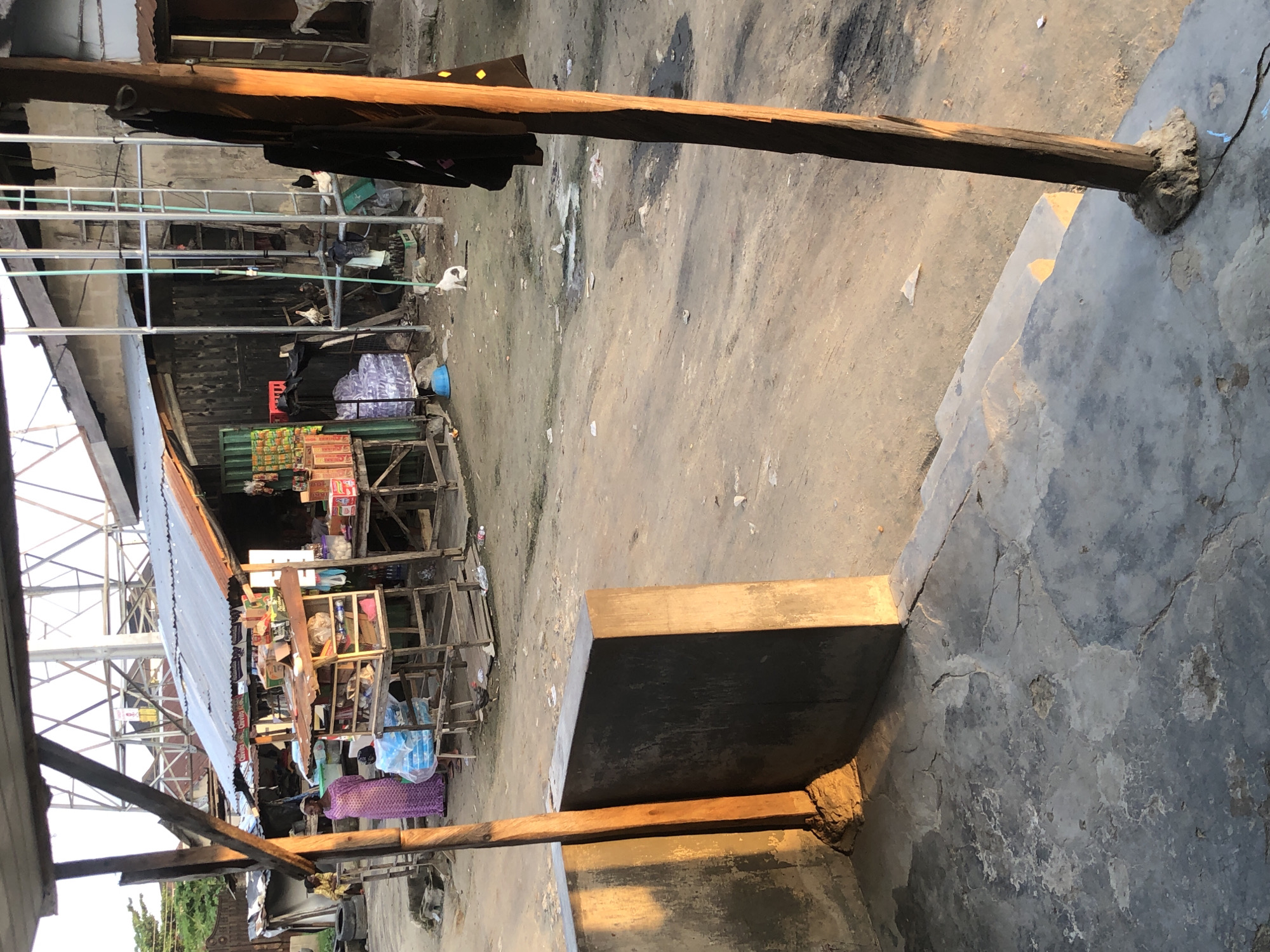 The image depicts an outdoor scene in Ado, Nigeria, characterized by an informal market and residential structures under a bright sky, likely during late afternoon due to the warm, directional sunlight.
In the immediate foreground, a cracked, uneven concrete floor leads to a pair of wooden support posts. The left post is integrated into a concrete ledge or wall, showing sunlit patches. The right post has black fabric draped over it.
The midground is dominated by a dirt ground with visible tire marks and scattered refuse including plastic bags and paper. Several makeshift market stalls, constructed from wood and corrugated metal sheeting, are positioned on the dirt. These stalls display various packaged goods, including food items and beverages. A woman in a long purple dress stands near one of the left-most stalls, while another individual is partially visible behind a stack of blue-wrapped items. Further right, a white cat with brown patches sits on the dirt, facing right. Several chickens roam near the stalls and closer to the background structures.
In the mid-to-background, various single-story buildings with corrugated metal roofs and unpainted, weathered walls are visible. A prominent, tall metallic structure, possibly a water tower or communication mast, with an open lattice design, rises above the buildings against the bright, slightly hazy sky. On the far right, a vertical metal scaffolding-like structure with green elements runs down its side. Sparse trees and foliage are visible on the far left.