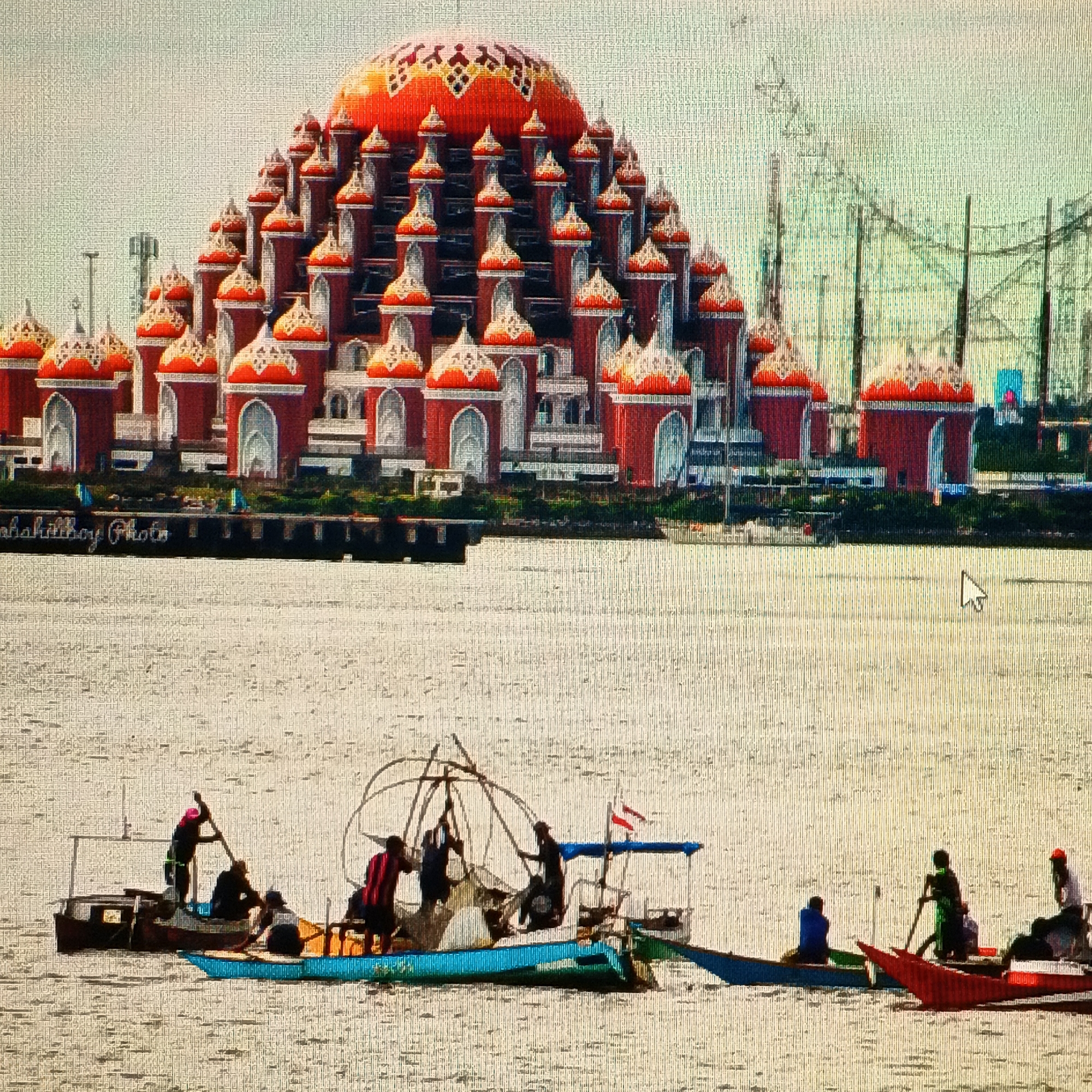 The image captures a wide view of a body of water, likely a harbor or bay, with a prominent building complex on the horizon and several boats with people on the water in the foreground.
The building complex, which appears to be the Mesjid Raya Baiturrahman in the city of Ende, Indonesia, dominates the upper half of the frame. It is characterized by a large central dome and numerous smaller domes, all adorned with vibrant orange and white patterns. The main structure is a rich reddish-brown color with white architectural accents. Behind the building, a series of tall, dark structures, possibly cranes or part of an industrial facility or theme park, are visible against a pale, hazy sky. A sign with faint white lettering is visible on the waterfront in front of the building, appearing to read "abahitboy Photo."
In the foreground, several traditional wooden boats are scattered across the water. One boat, positioned slightly to the left, has at least three individuals on board; one person stands at the stern and appears to be steering or rowing with a long pole, while two others are seated. This boat has a light-colored deck and a dark hull. Next to it is a longer, blue boat with at least two people, one of whom is actively rowing with a paddle. Further to the right, a larger boat with a rudimentary shelter is visible. This boat carries multiple individuals, some of whom appear to be tending to fishing nets or equipment. A small red and white flag flies from a mast on this boat. To the far right, another boat, this one red, is partially visible with at least two figures on board. The water is calm and appears somewhat choppy, reflecting the light. The overall impression is a juxtaposition of a grand architectural landmark with the everyday activities of local fishermen on the water.
