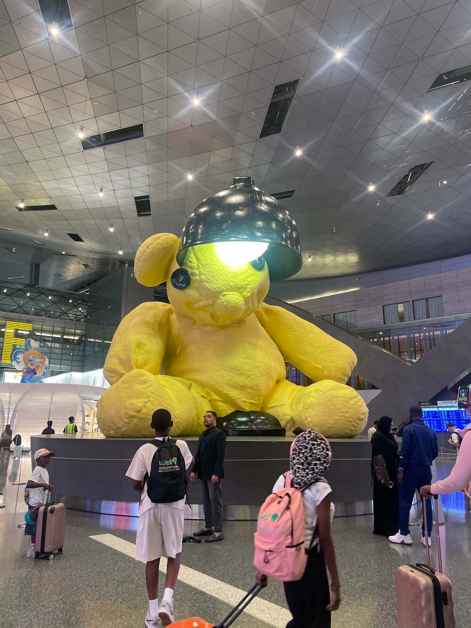 The image shows a large yellow teddy bear sculpture with a black lampshade on its head, illuminated from within, located in the interior of Hamad International Airport in Doha, Qatar. The sculpture is the centerpiece of the main concourse, which features a modern, expansive ceiling with triangular panels and recessed lighting. Several people are visible in the foreground and background, including children with suitcases and adults dressed in casual and formal attire. The environment appears to be a busy airport terminal, with escalators and signage visible in the distance. The lighting is a mix of artificial light from the ceiling and the sculpture itself, creating a bright and somewhat dramatic atmosphere.