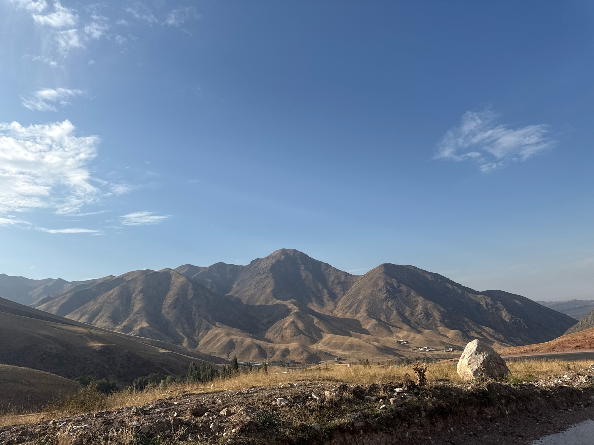 This image depicts a vast, arid mountain landscape under a clear blue sky with scattered wispy clouds. The mountains, characterized by their dry, barren slopes, are sculpted by erosion, revealing a network of valleys and ridges. Patches of dry grass and sparse vegetation dot the lower hills. In the foreground, a rough, rocky terrain with dry grasses leads to a large, light-colored boulder marked with red symbols. A paved road is visible on the right side of the image, curving away into the distance. In the middle ground, a small cluster of buildings, possibly a village, is nestled among the hills. The overall scene conveys a sense of rugged natural beauty and isolation in a mountainous region. The location is specified as Tatyr, Kyrgyzstan.