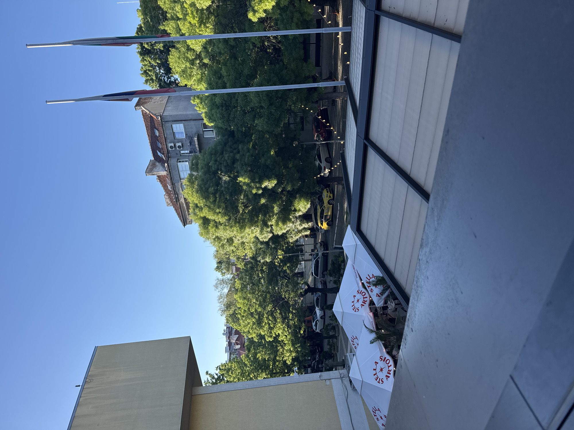 The image shows a view from an elevated position in Sofia, Bulgaria. Two flagpoles, bearing Bulgarian flags, stand in the background near a building with a red-tiled roof and cream colored facade. In the mid-ground, there are green trees lining a street with several parked vehicles. Below the vantage point, there are several white Stella Artois-branded umbrellas covering a patio. The lighting suggests it is daytime.