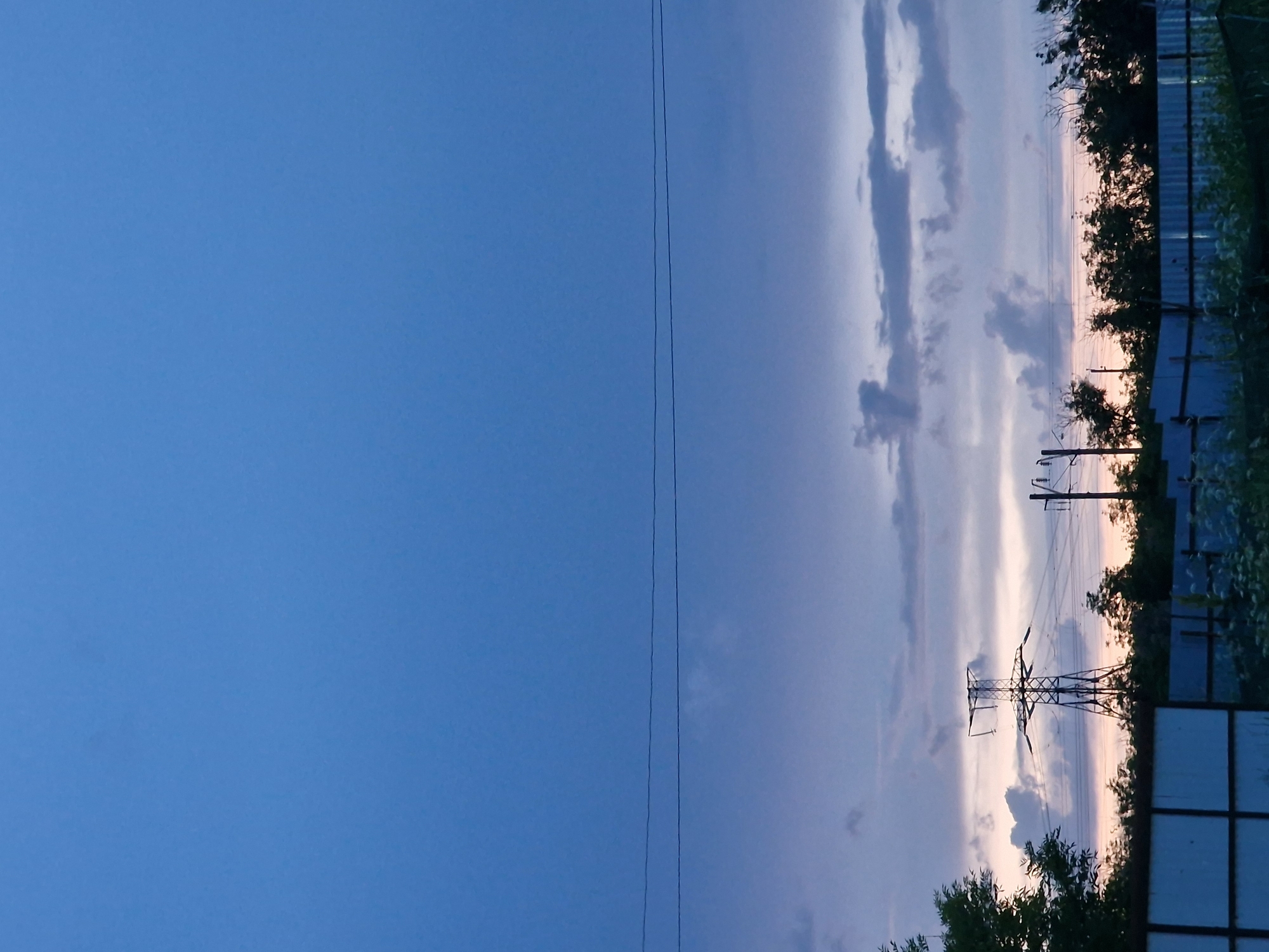 The image depicts an industrial landscape at dusk, likely in or near Kursavka, Russia. A prominent lattice tower, characteristic of an electrical pylon, stands silhouetted against a twilight sky. The sky is a gradient of deep blue at the top, transitioning to lighter blues and purples with streaks of pale orange near the horizon, indicating the setting or rising sun. wispy clouds are visible, scattered across the mid-sky.
In the foreground, a light blue corrugated metal fence stretches horizontally across the frame, partially obscuring the scene behind it. Above and behind the fence, a dense line of dark green trees forms a natural barrier. To the left of the pylon, the dark silhouette of a large tree with dense foliage is visible. To the right of the pylon, another utility pole with insulators can be seen. The ground beyond the fence appears to be covered in sparse vegetation, with some lighter colored plants or flowers visible. Two thin, dark electrical wires are strung across the upper portion of the frame, parallel to each other and stretching from left to right. No people are visible in the image. The overall atmosphere is subdued and industrial, characteristic of a rural or semi-urban area during twilight.