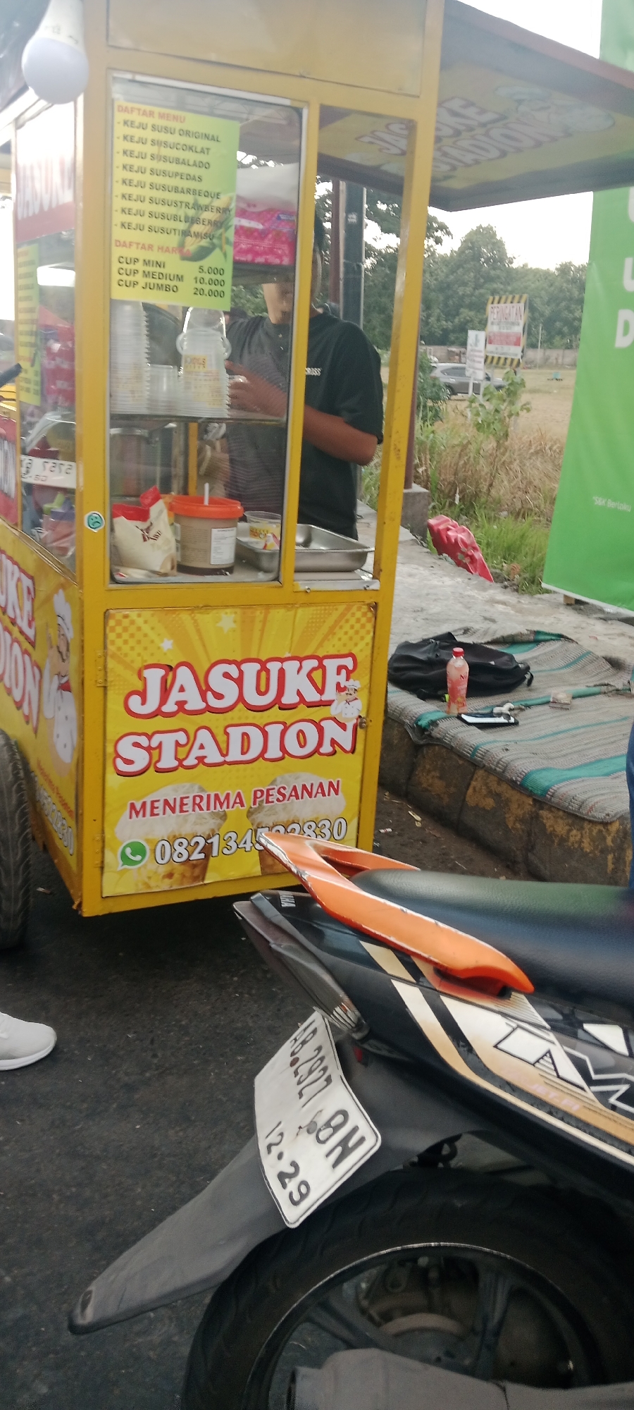 This is a medium-angle, ground-level shot capturing a street food cart. The cart is primarily yellow and features a menu board and branding that reads "JASUKE STADION." The menu board lists various flavors of cheese milk and prices for different cup sizes. A person, likely the vendor, is partially visible behind the glass enclosure of the cart. In the foreground, a motorcycle is parked, partially obscuring the cart's lower section. The motorcycle has a license plate visible with the characters "AB. 2927 8N" and "12. 29". The scene appears to be outdoors on a paved surface, with dried grass and trees visible in the background. The lighting suggests it is daytime. The presence of a street food cart and motorcycle indicates a public or semi-public area within the city of Ngemplak, Indonesia, likely a roadside or market setting.