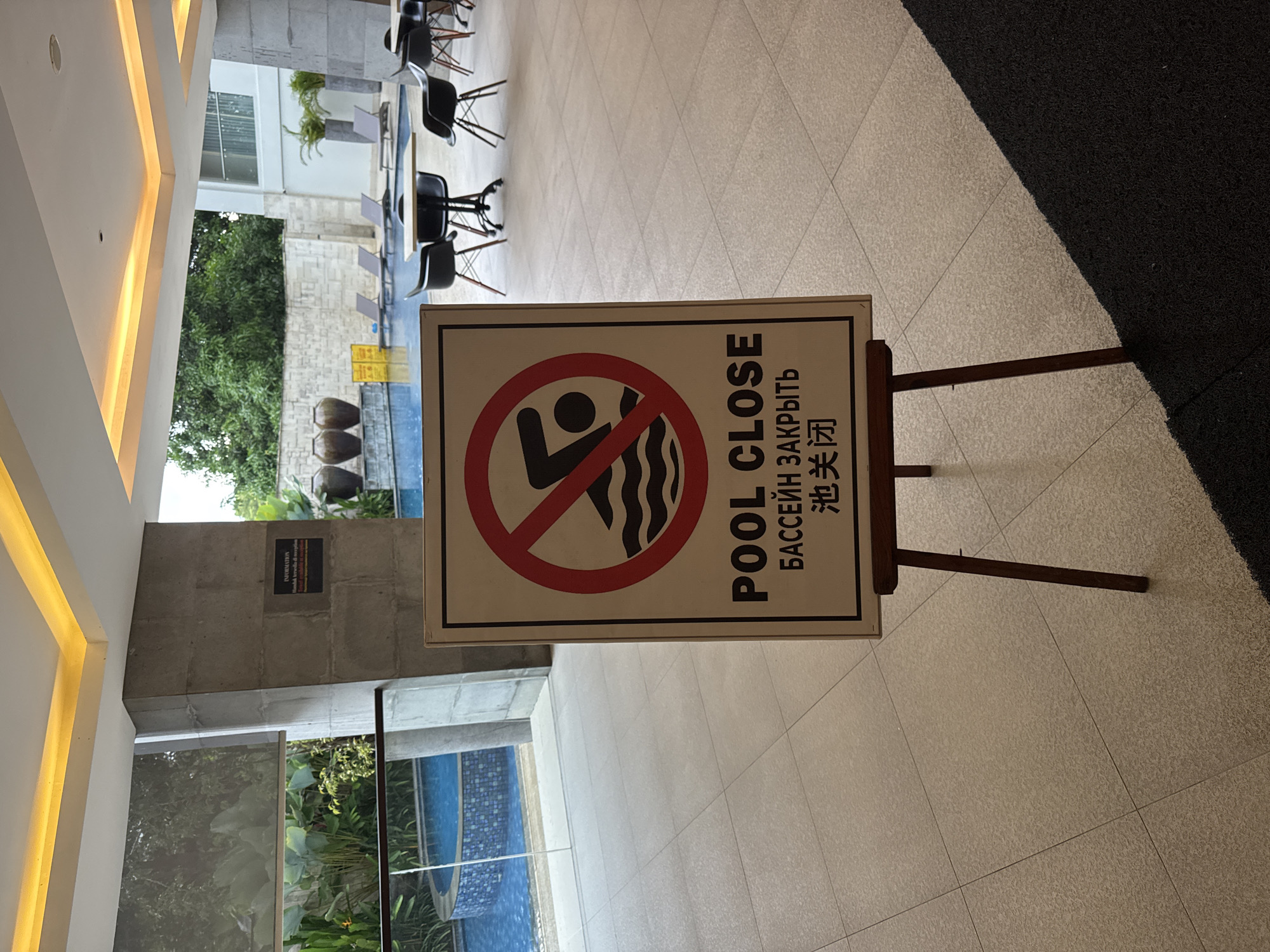 The image displays an indoor-outdoor transitional area, likely within a resort or hotel in Kuta Selatan, Indonesia. The central focus is a white rectangular sign, propped on a dark wooden easel, indicating that the pool is closed. The sign features a universally recognized "no swimming" symbol – a black silhouette of a person in water with a red circle and diagonal line through it. The text "POOL CLOSE" is written prominently in English, followed by Russian ("БАССЕЙН ЗАКРЫТЬ") and Chinese ("泳池关闭").
The scene is brightly lit, suggesting daytime. The flooring consists of light-colored square tiles, leading towards a swimming pool with bright blue water, partially visible beyond a glass barrier. Lush green tropical foliage and trees are abundant in the background, hinting at a warm climate. Several black tables and chairs with slender legs are arranged on the tiled patio area, providing seating for guests. Large, dark decorative pots are situated near the edge of the pool.
On a concrete wall in the background, a smaller, dark rectangular sign with white text is visible, containing the word "INFORMATION" and some Indonesian text, including "Dilarang membawa..." (meaning "Forbidden to bring..."), though the rest is unreadable due to the angle and rotation. Architectural elements include a light-colored ceiling with illuminated recesses and textured concrete walls. A dark grey rug or mat is visible on the right side of the image, contrasting with the tiled floor. No people are present, giving the area a quiet and unoccupied appearance.