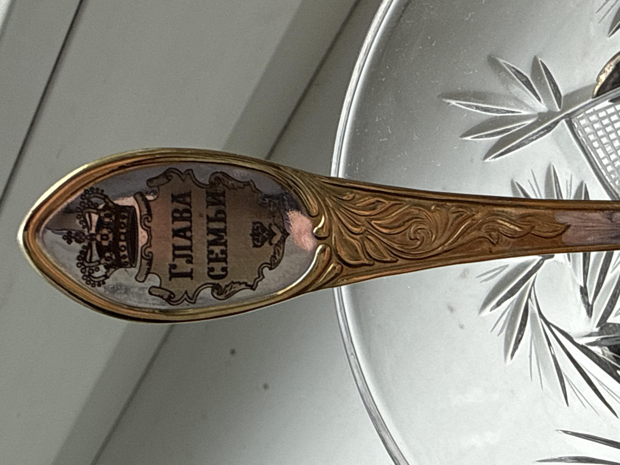 This is a close-up, rotated shot of an ornate spoon with decorative elements. The spoon's handle has a gold-colored, embossed floral design. The bowl of the spoon is oval-shaped and appears to be made of silver or a similar metal, bordered by gold. Inside the bowl, there's an inscription in Cyrillic script within a decorative cartouche. The text reads "ГЛАВА СЕМЬИ," which translates to "Head of the Family." Above the text, there is a detailed depiction of a crown. The spoon is positioned over a piece of clear, cut glass, possibly a bowl or plate, with a starburst or fan pattern etched into it. The background is a plain, light-colored surface, suggesting an indoor setting with diffused lighting. There is no indication of the time of day, weather, or a specific geographical location. The image focuses solely on the spoon and the glass object it rests upon, with no people or specific activity visible.