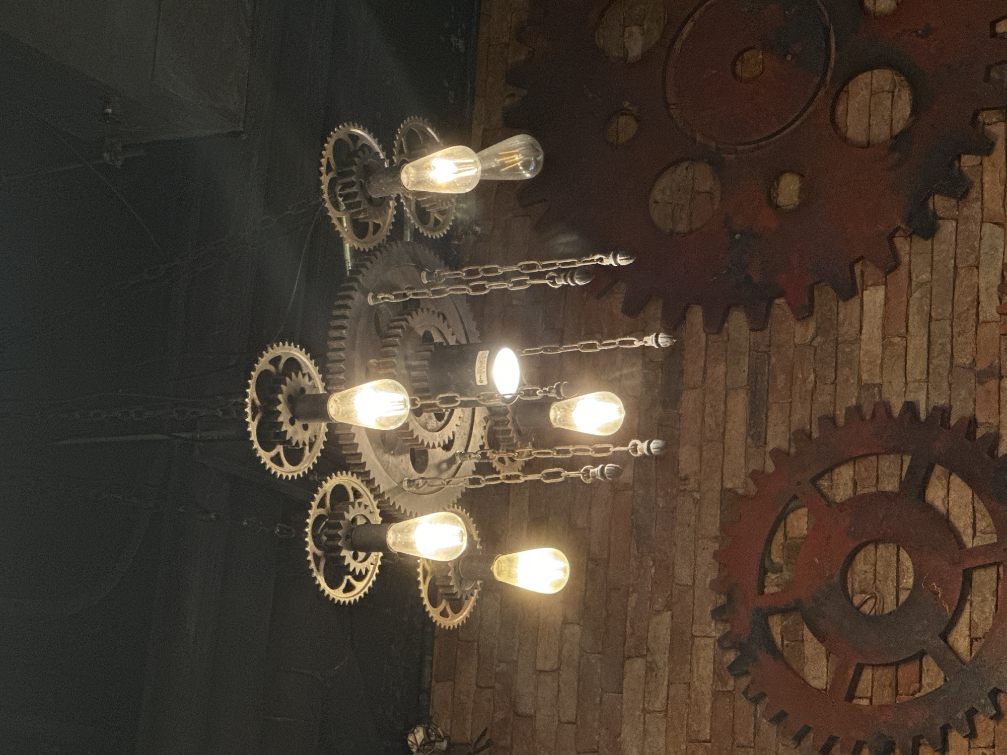 The image shows a steampunk-themed light fixture attached to a brick wall. The light fixture is composed of metal gears and exposed light bulbs. The brick wall behind the fixture is rough in texture. The image was taken indoors. There is no information available from this image regarding the location within Bac Nha Trang Ward, Vietnam. No people are visible.