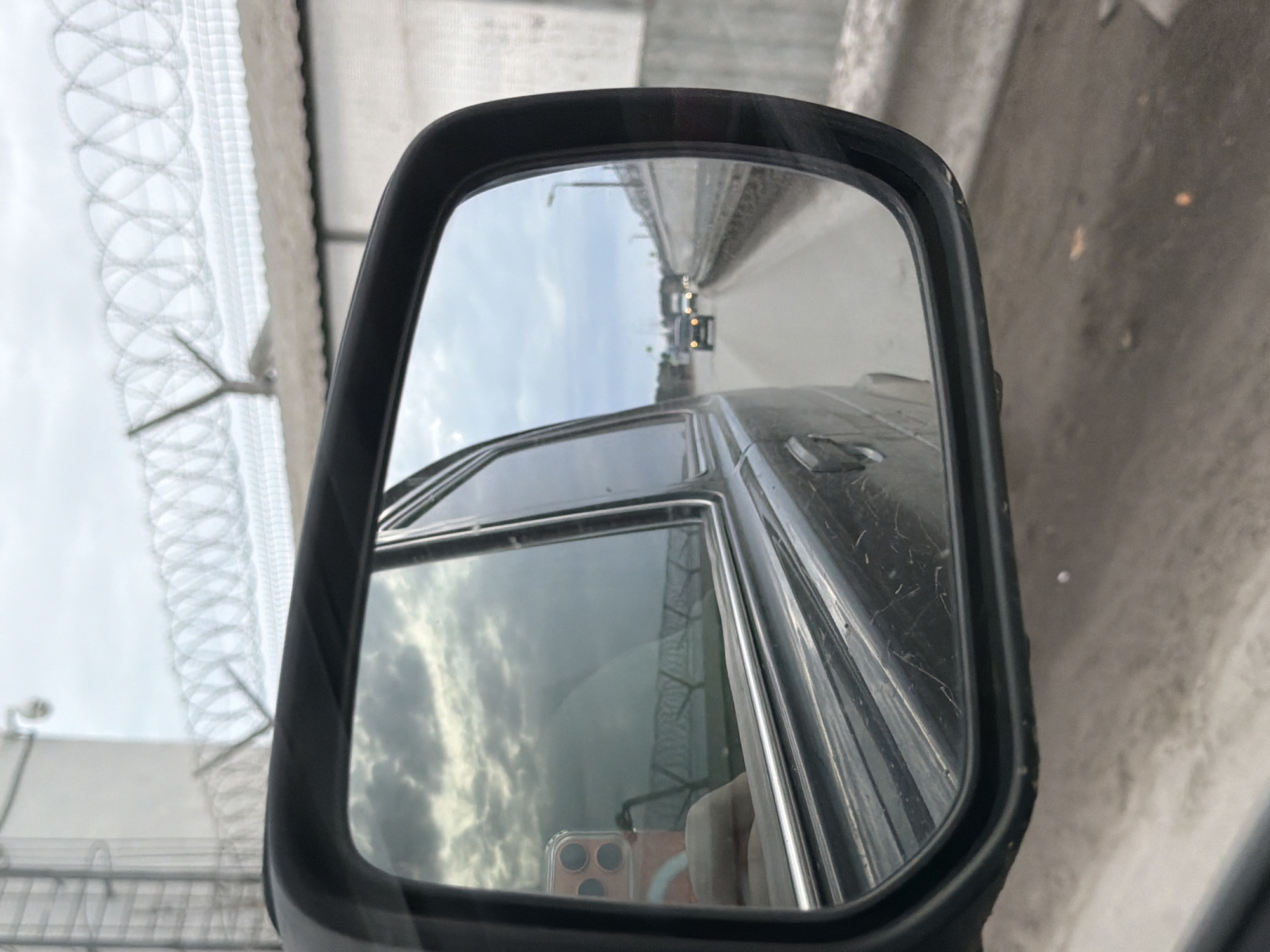 The image shows a driver-side rearview mirror of a vehicle. The mirror reflects a road with other vehicles and a highway. The image also contains a glimpse of a razor wire fence through the window. The reflection in the side window shows a phone. The location is most likely Chaikovsky, Russia, due to the prompt’s specification.