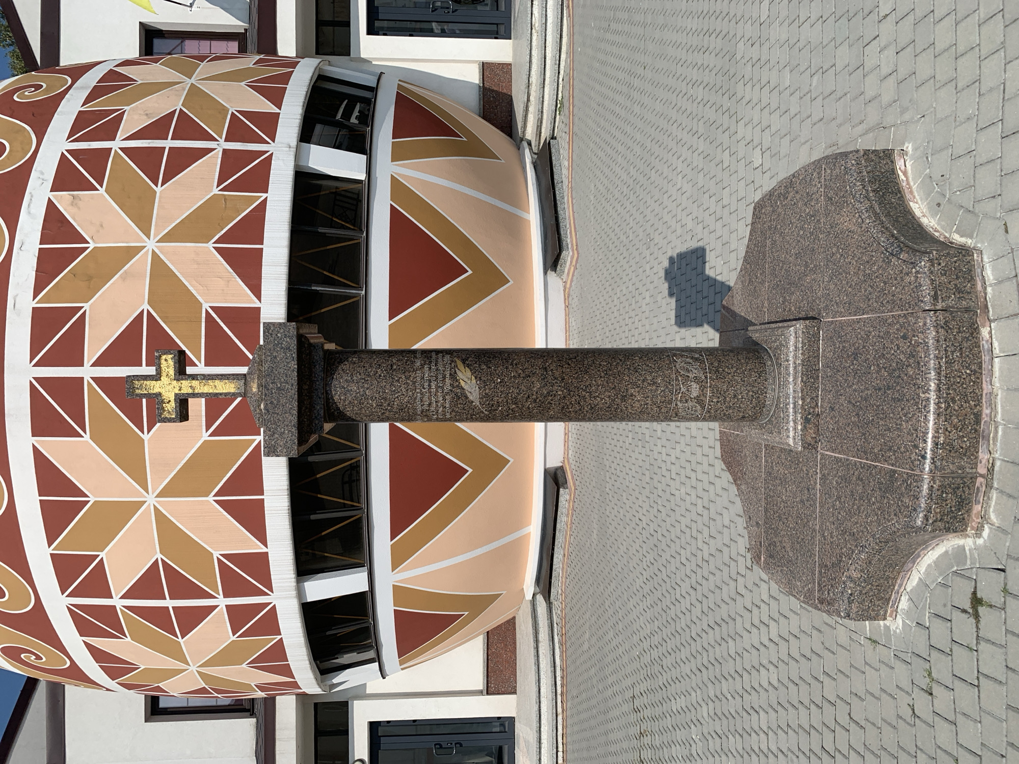 The image depicts the exterior of the Pysanka Museum in Kolomyia, Ukraine. In the foreground, a tall, cylindrical granite monument stands centrally on a multi-tiered, octagonal granite base. A small, golden cross is affixed to the top of the monument. The monument is inscribed with text and a feather motif, though the text is largely illegible in the image. The monument casts a distinct shadow on the grey, interlocking paver walkway that surrounds it. Behind the monument is the museum building, a large, egg-shaped structure adorned with a colorful geometric pattern of red, brown, and light orange triangular shapes and star-like designs. The museum has a white facade and several windows with dark frames. The sky above is clear and blue. The overall scene is that of an outdoor public space in front of a cultural landmark.