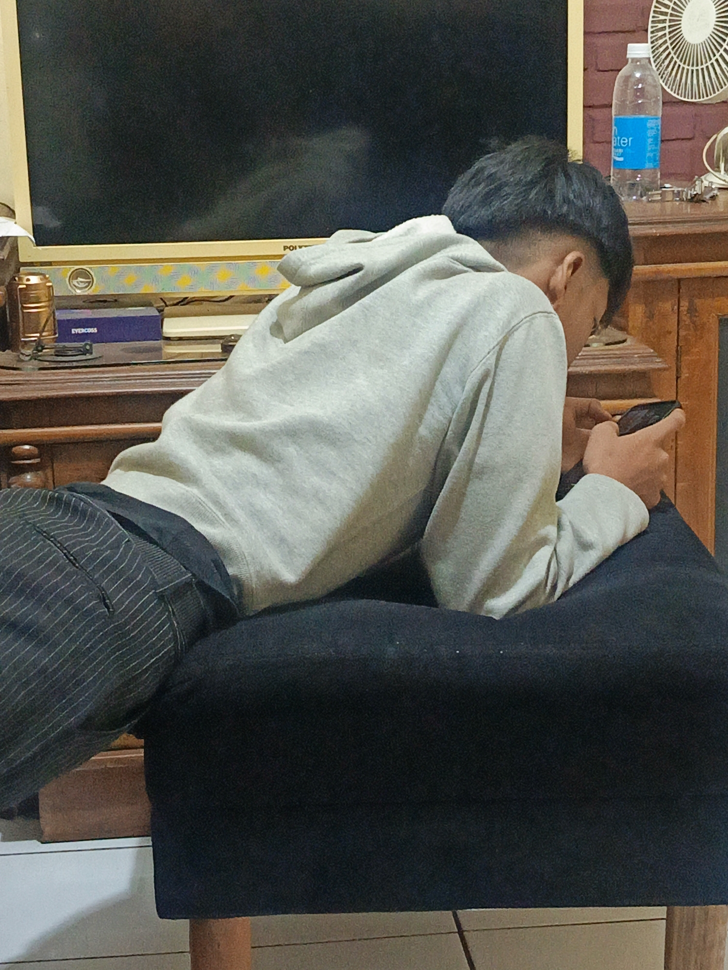 A young man with short, dark hair, wearing a light gray hooded sweatshirt and dark pinstriped pants, is lying on his stomach on a dark gray cushioned surface. His right arm is extended, and he is holding a black smartphone in his right hand, looking down at it. The background shows a television set with a beige frame, a wooden cabinet, and a bottle of water on a shelf. The scene appears to be indoors, likely in a residential setting in Margajaya, Indonesia, given the context.