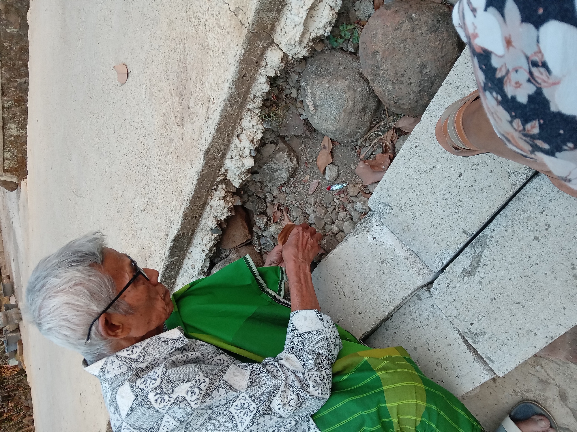 An elderly person with gray hair and glasses is crouching on concrete blocks. They are wearing a white patterned shirt and a green sarong. The person is holding a small object in their hands. The environment appears to be outdoors, with rough ground, rocks, and dried leaves visible. The image is taken from a high angle, looking down at the person and the surrounding area. The location is identified as Kedunggudel, Indonesia.