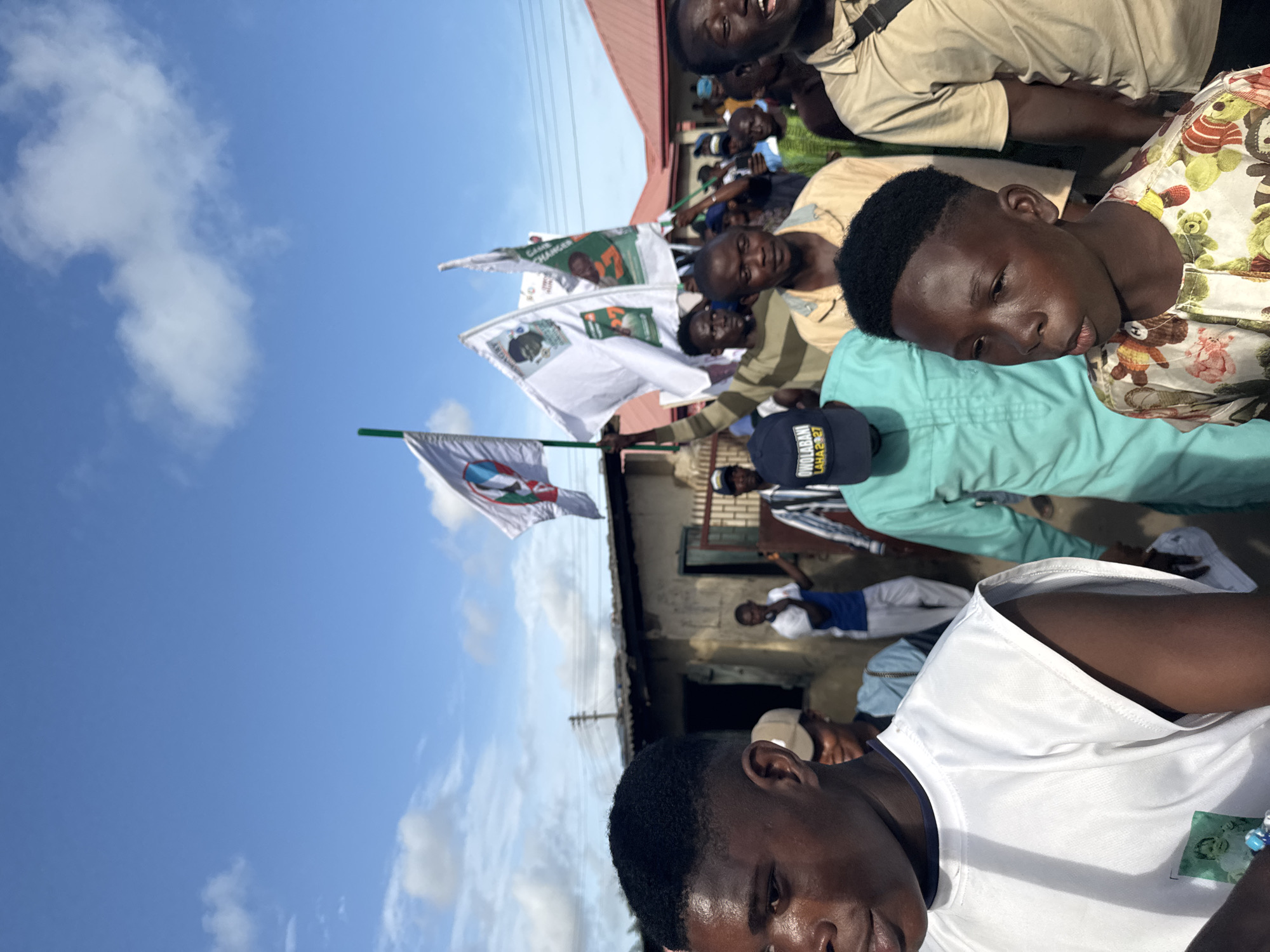 The image depicts a crowd of people in Badagry, Nigeria. Some individuals are holding political flags. The flags are white with green and blue elements. One flag contains a portrait of a man. The sky is blue with scattered clouds. A building with a red roof is partially visible in the background. Some individuals are wearing casual attire, including white shirts, green shirts, and a hat with text.