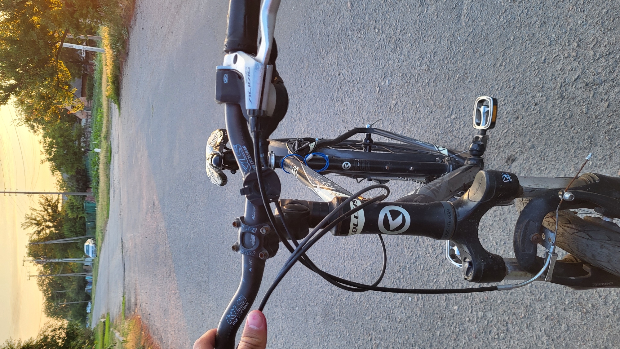 A first-person perspective of a bicycle in motion on an asphalt road. The handlebars, with KLS Rise Crossbar branding, are visible in the foreground, along with brake levers and cables. The front fork and part of the frame are also in view. The road ahead shows houses and trees lining the sides, with a car parked in the distance. The sky suggests it is late afternoon or early evening, with a warm, golden light. The environment appears to be a rural or suburban area.