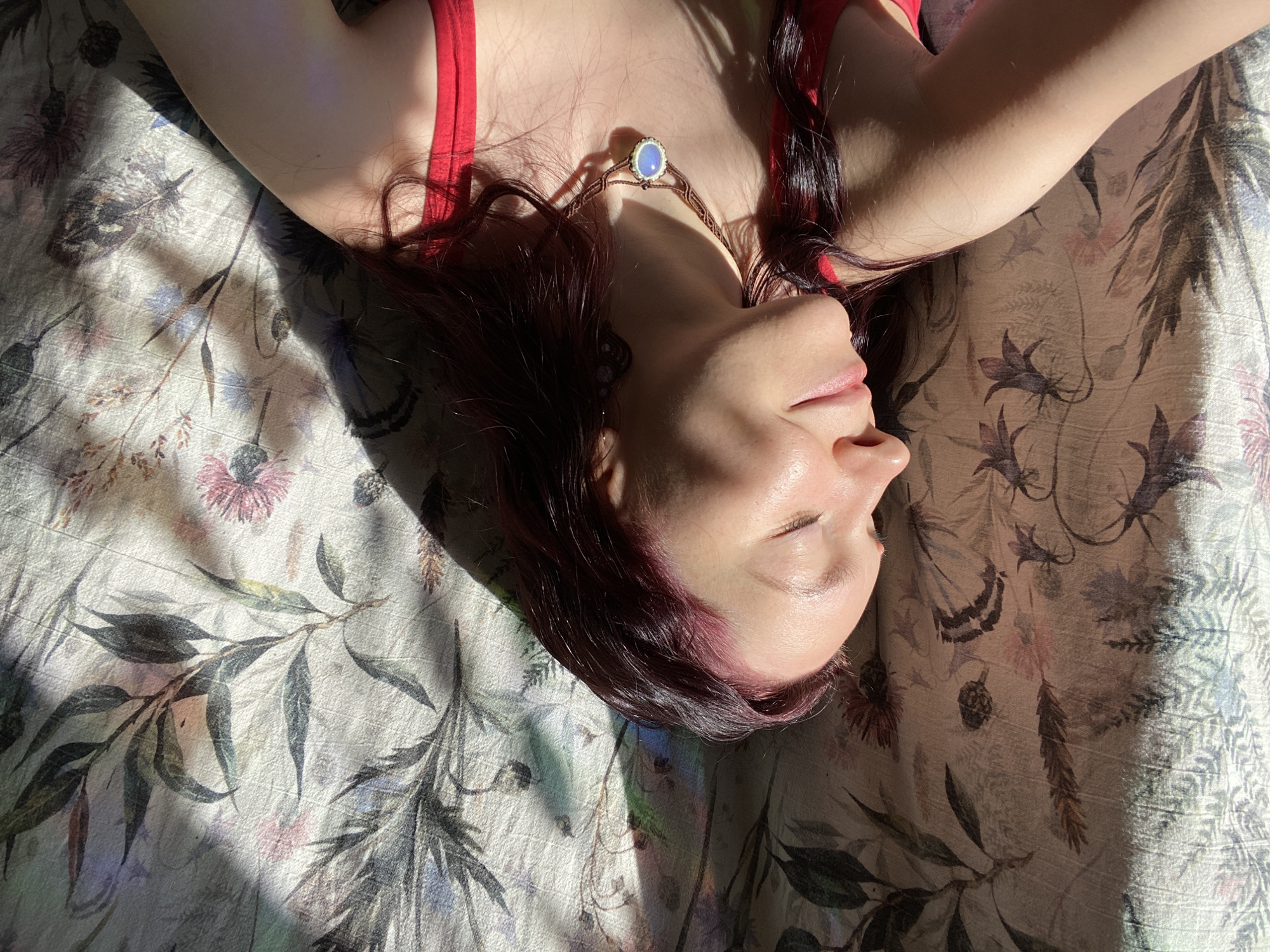 A woman with reddish-purple hair is lying on a floral-patterned surface in Chaikovsky, Russia. She is wearing a red top and a necklace with a blue stone pendant. Light and shadows play across her face and the surface beneath her. The floral pattern consists of various plants and flowers, including leaves, branches, and blooms, with a color palette of muted grays, blues, and pinks on a light background. The image captures a moment of relaxation or rest.