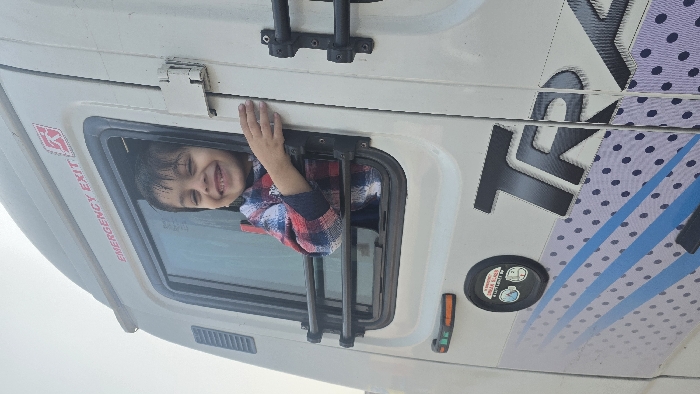 A smiling child in a red and blue plaid shirt peeks out from a rectangular window with an "EMERGENCY EXIT" sign. The window is part of a white vehicle with black fixtures and a ladder attachment above. The vehicle has the word "TRUCK" in large black letters on its side, along with a blue striped pattern and black polka dots.