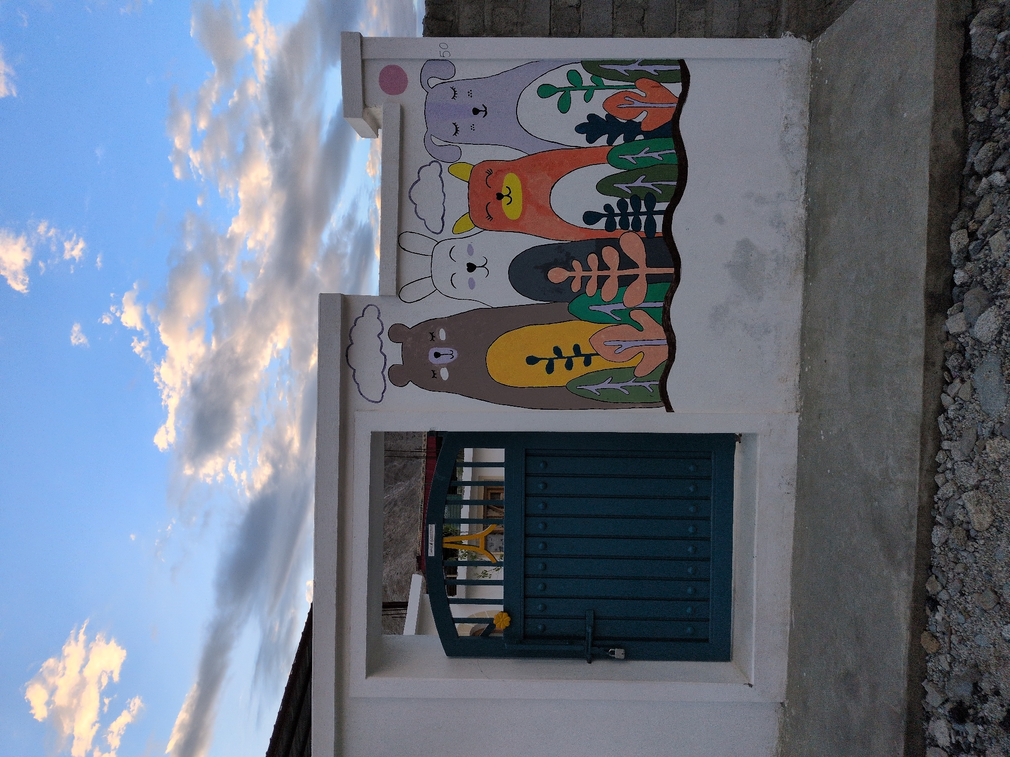 The image shows a white wall with a mural of four cartoon animals: a brown bear, a white rabbit, an orange fox, and a purple dog. They are depicted with closed eyes and smiling faces, standing behind stylized trees and plants. The mural is on the side of a building in Leh, India, where the sky is blue with scattered clouds. A dark teal metal gate with a padlock is visible on the left side of the wall. The ground in front of the wall is concrete, with gravel and rocks at the base.