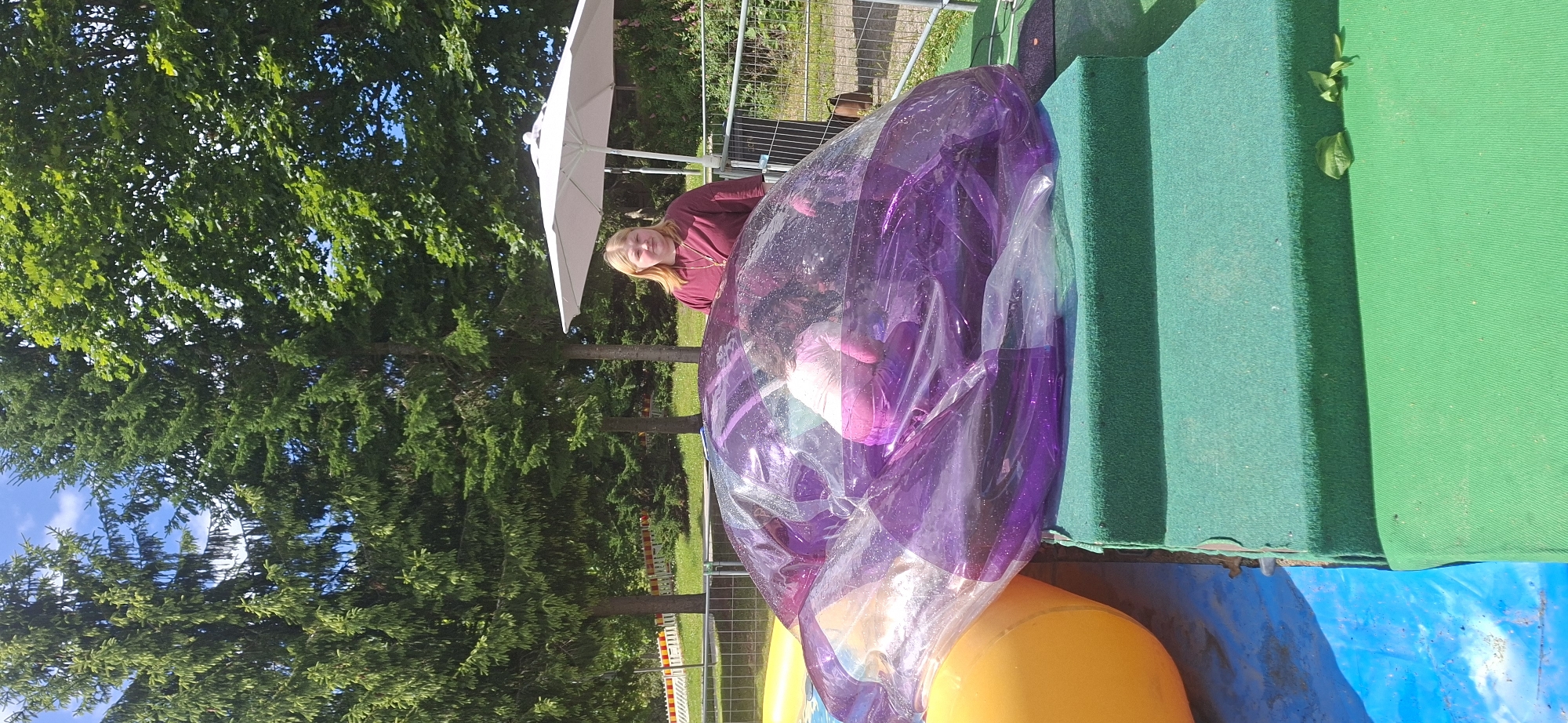 An outdoor scene in Lahti, Finland features a large, transparent purple inflatable ball with a person inside. A woman stands behind the ball, looking at the camera. The inflatable is on green steps and a blue mat. Trees are in the background, along with a white umbrella and a metal fence. The sky is blue with some clouds.