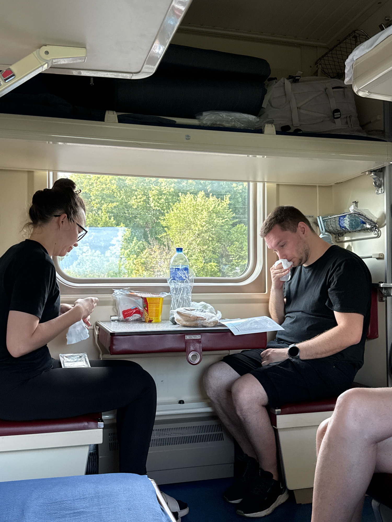 A medium shot captures two individuals seated inside a train compartment. A woman with her hair pulled up in a bun, wearing glasses and a black t-shirt, is on the left. She is holding a white napkin in her hands and appears to be preparing something from a small foil container. To her right, a man wearing a black t-shirt and black shorts is seated. He is holding a white napkin to his nose and appears to be blowing his nose. He is wearing a watch on his left wrist. In front of them is a small table with various items, including a bottle of water, a box of snacks, and some bread. The compartment has overhead storage shelves filled with luggage and personal items, typical of a train journey in Rostov-on-Don, Russia. The window behind them shows a blurred view of green trees and foliage, suggesting the train is in transit, possibly on the outskirts of the city. The seating appears to be upholstered in a dark red fabric.