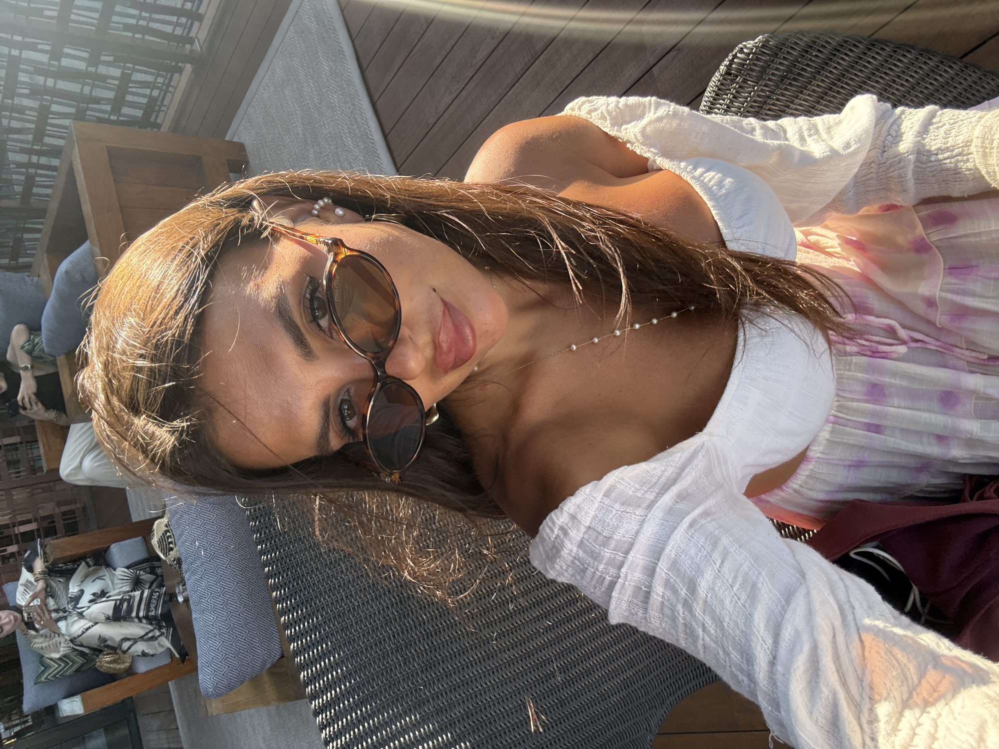 This is a close-up, selfie-style image of a woman with long brown hair, wearing sunglasses and an off-the-shoulder white top. She is seated outdoors, with a wicker chair visible in the foreground. In the background, other individuals are seated in chairs on a wooden deck. The scene suggests a leisurely outdoor setting, potentially a resort or private residence. The lighting indicates it is daytime, with sunlight casting shadows. The overall impression is of a relaxed, warm-weather environment. The provided crop hints at a location in Bodrum, Turkey, a coastal city known for its resorts and tourist attractions.