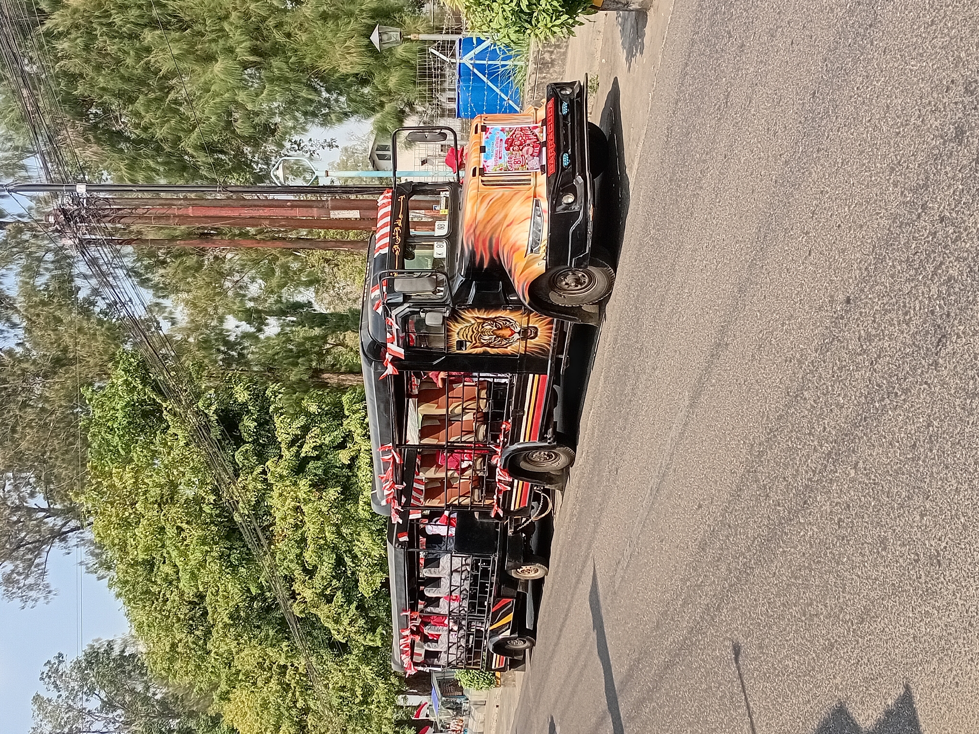 A black and orange converted truck with open-air seating, resembling a bus, is parked on a paved road in Gebog, Indonesia. The vehicle is decorated with red and white flags and has a large tiger graphic on its side. Passengers are seated inside. Trees line the road, and utility poles with numerous wires are visible in the background. The scene appears to be during daylight hours.