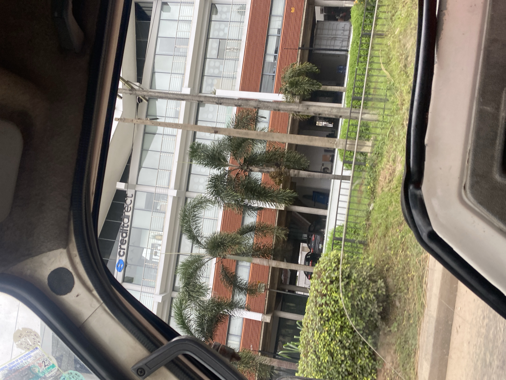 In Ikeja, Nigeria, a modern multi-story building with a distinctive facade of horizontal orange louvers and large glass windows stands prominently. The building's exterior is sleek, with white balconies and railings. In front of the building, several palm trees with feathery fronds are planted, adding a tropical element to the urban landscape. A low green hedge and a metal fence line the perimeter. The view is from inside a vehicle, looking out through the windshield and window frame. A black car is visible parked beneath the covered section of the building, along with other parked vehicles. The sky is overcast, suggesting a cloudy day.