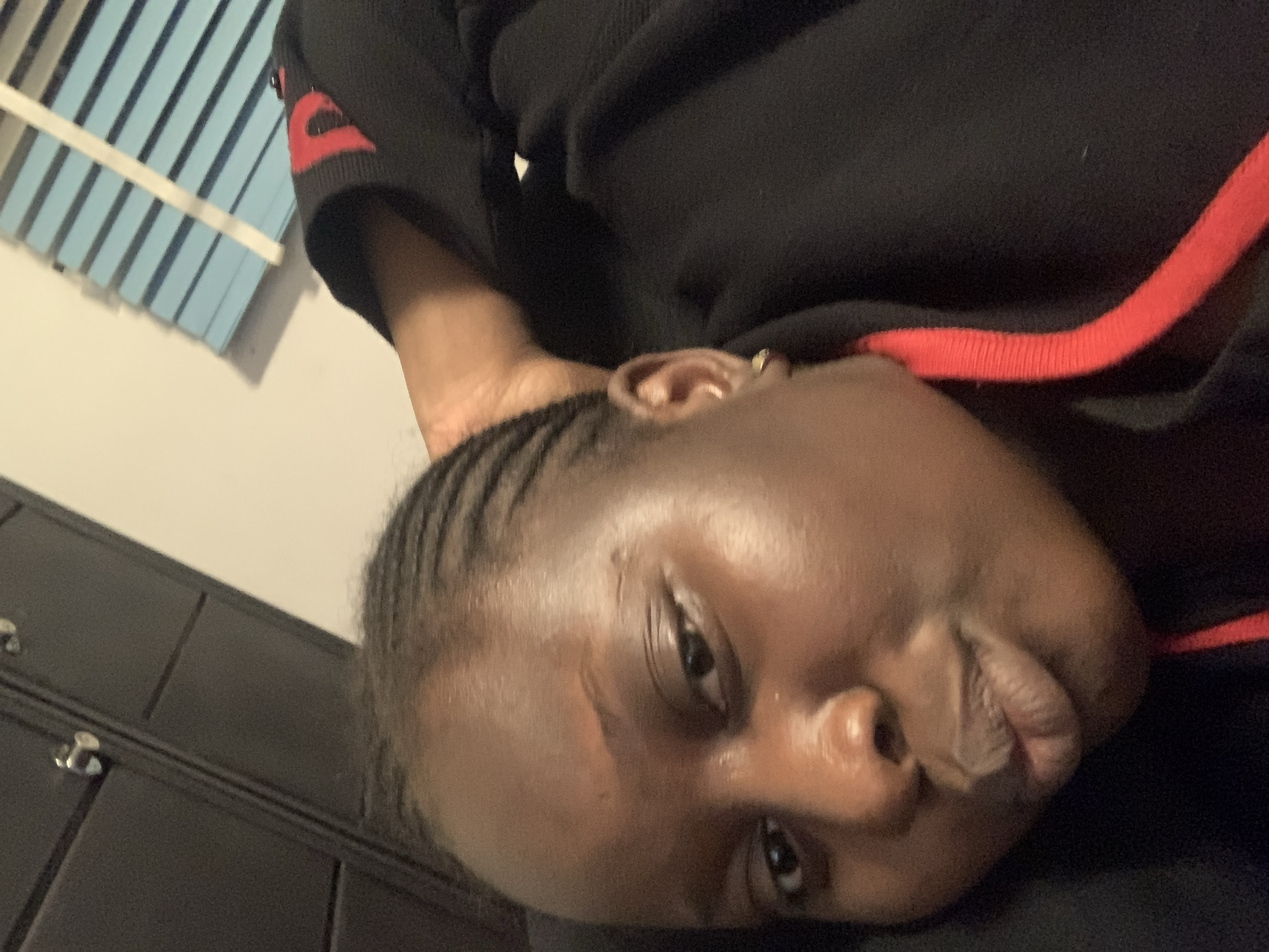 A young woman is captured in a close-up selfie, her face angled slightly towards the camera. She appears to be indoors, with a dark closet or cabinet visible in the upper left corner of the frame. The background includes a wall with horizontal blue blinds. The woman has dark skin and her hair is styled in braids. She is wearing a black garment with a red trim. Her right arm is bent and resting behind her head. Her lips are slightly pursed.