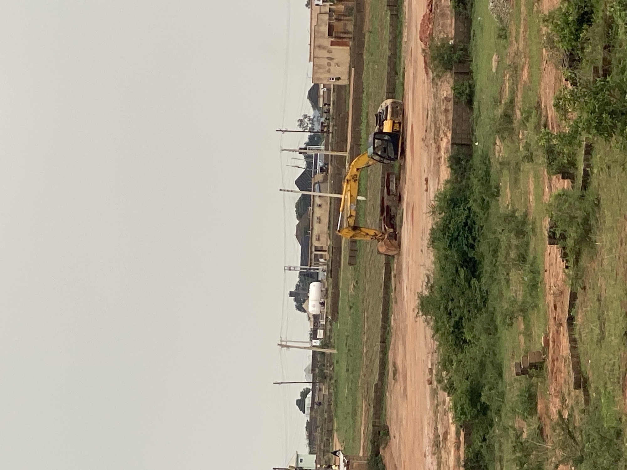 The image captures an outdoor scene in Jos, Nigeria, depicting a construction site under an overcast sky. In the foreground, a dirt patch leads to a grassy area with some scattered bricks and greenery. A yellow excavator is parked on the dirt, appearing to be part of ongoing construction work. In the middle ground, there are more grassy areas and several low-lying structures, possibly workshops or storage units, constructed with mud bricks or concrete. Further in the background, a large white cylindrical tank, likely for storage, is visible, along with several buildings with pitched roofs and utility poles with power lines stretching across the landscape. A few individuals are present, scattered across the site, some appearing to be workers. The overall impression is that of a developing area or an industrial zone within Jos.