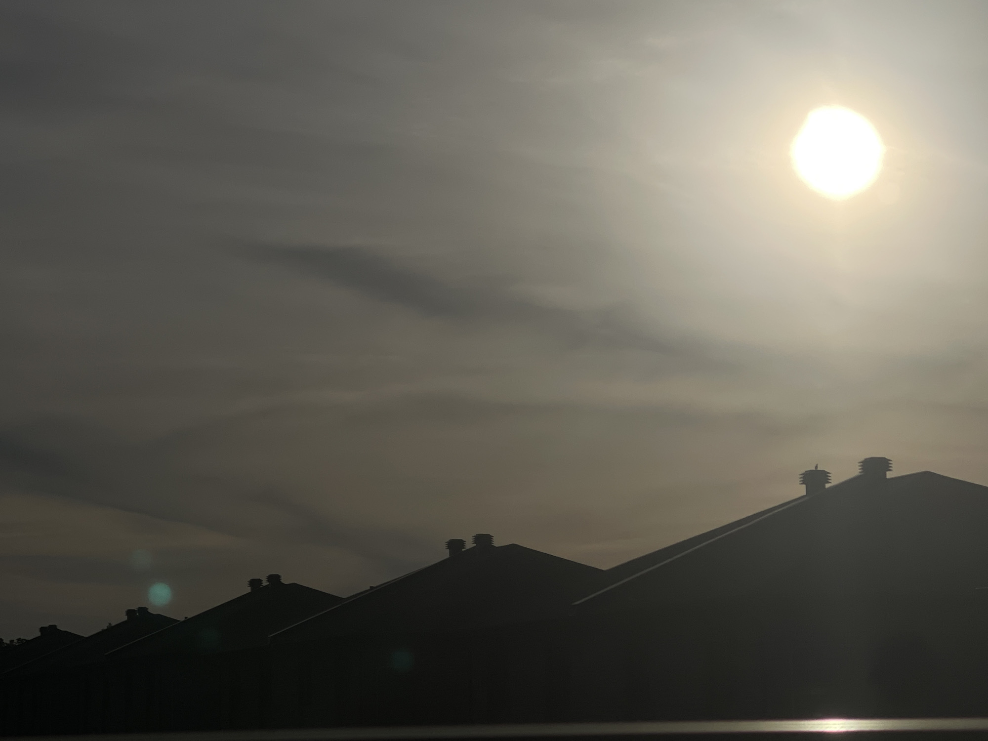 The image presents a dramatic scene of silhouetted rooftops against a luminous, partly cloudy sky, set in Gatineau, Canada. The bright, almost blinding sun is prominently featured in the upper right quadrant, emitting a powerful glow that illuminates the surrounding sky in shades of warm white and hazy yellow. The sky transitions from a darker grey on the left to a brilliant luminosity around the sun, with discernible streaks of clouds adding texture and depth.
In the foreground and midground, the angular forms of several building roofs are silhouetted, appearing as dark, unidentifiable shapes against the intense brightness of the sky. These structures feature distinct ventilation pipes or small chimneys protruding from their ridges, with a tiny, unidentifiable object perched on one of them in the mid-right.
Notable details include prominent lens flares, specifically a greenish-blue one in the lower-left portion of the frame, and a bright, elongated reflection along the very bottom edge, possibly from a reflective surface like water or glass. The stark contrast between the dark foreground and the brilliant sky strongly suggests either early morning or late afternoon. The overall atmosphere is serene and natural, with no people or visible text present.