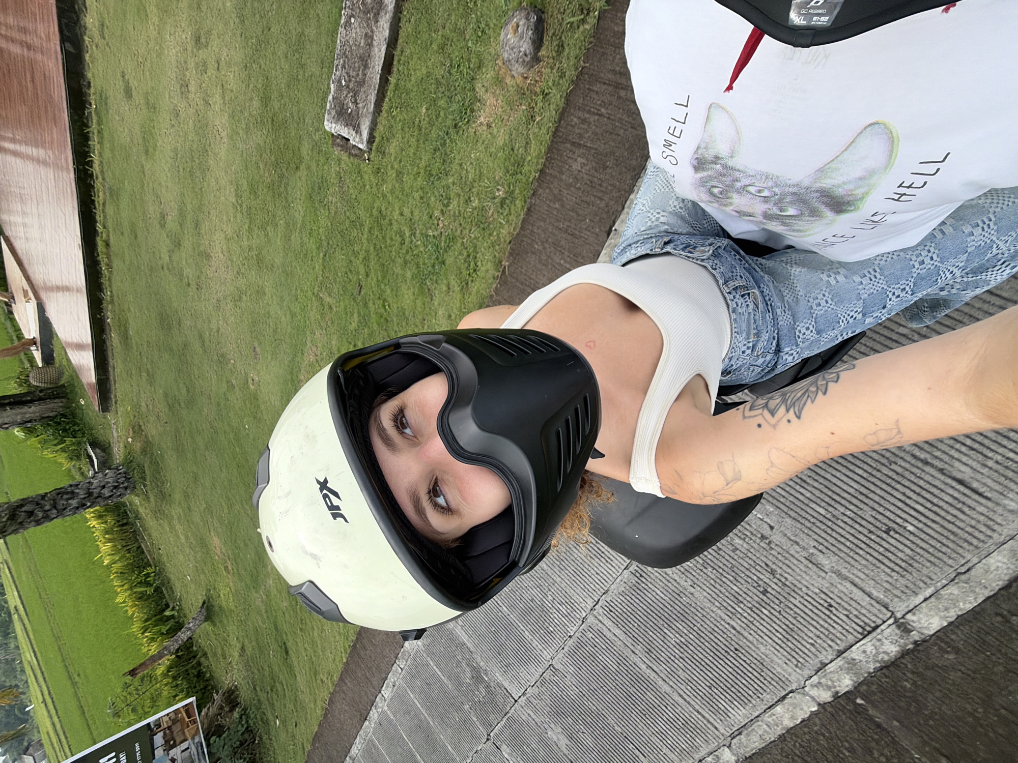 A first-person perspective, overhead shot shows a young woman wearing a cream-colored motorcycle helmet with a black visor, white tank top, and light blue patterned jeans. She is looking up and to her left with a slight smile. Her right arm, visible in the foreground, has several tattoos. She is seated on the back of a motorcycle. To her right, a white t-shirt with a graphic print and text is visible. The background shows a paved walkway next to a grassy area and a rice paddy field. The scene appears to be in a tropical location, consistent with North Kuta, Indonesia, given the lush green vegetation and rice fields. The lighting suggests it is daytime.