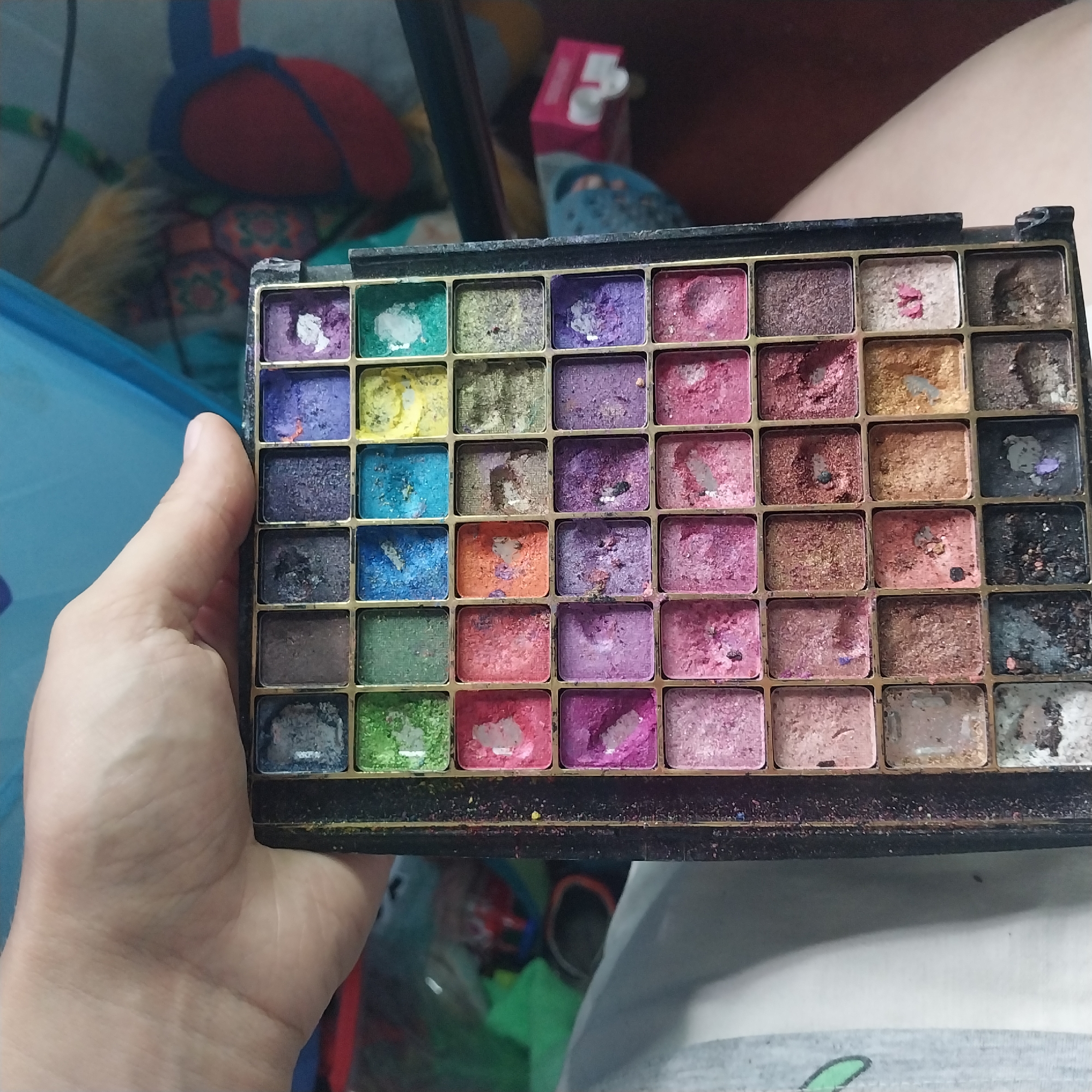 An overhead, close-up shot shows a person's hand holding an open eyeshadow palette. The palette is rectangular with a black exterior and a grid of 40 eyeshadow pans. The pans are a mix of matte and shimmer finishes and include shades of pink, purple, blue, green, orange, brown, and gray. Many of the eyeshadows appear well-used and have visible indentations from application. The background is out of focus but appears to be an indoor setting with a blue surface and some colorful objects. The lighting is neutral, and there are no discernible landmarks or elements that would indicate a specific city or location.