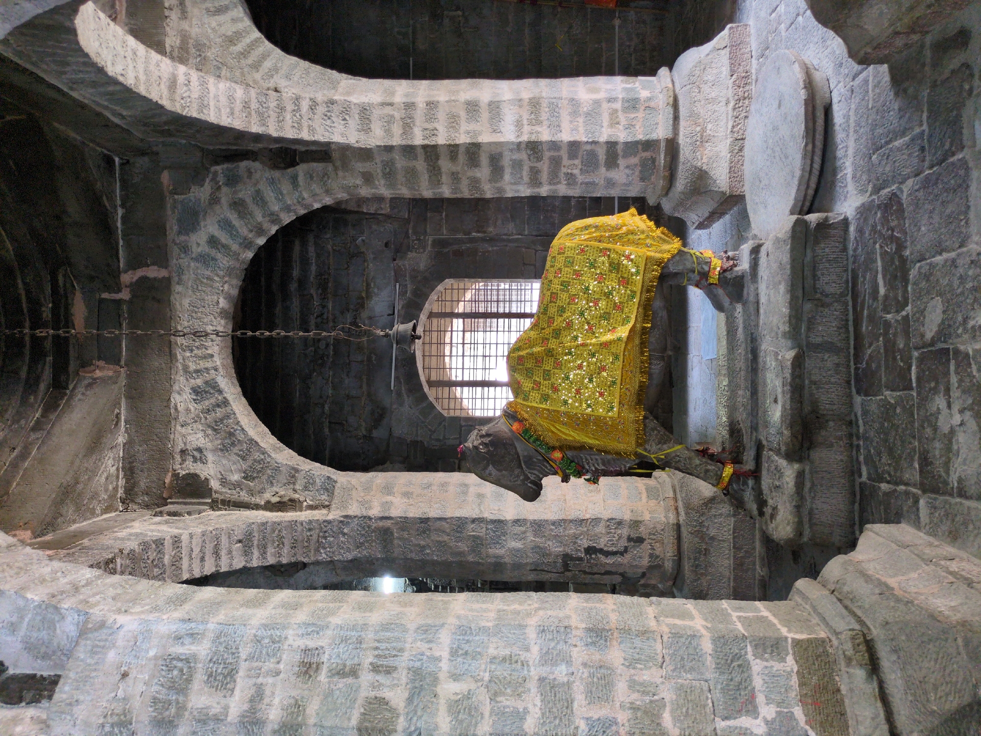 The image shows the interior of a stone structure, possibly a temple, with large, rough-hewn stone pillars supporting arched ceilings. In the center foreground, a stone sculpture of a bull stands on a tiered stone platform. The bull is draped with a yellow cloth adorned with intricate golden patterns, including swastikas. The bull also wears a garland of flowers around its neck. Behind the bull, a large arched window with a metal grate allows light to enter the space. A chain hangs from the ceiling, suspending a small bell above the bull. The floor is paved with flagstones. The overall impression is one of ancient architecture and religious iconography.