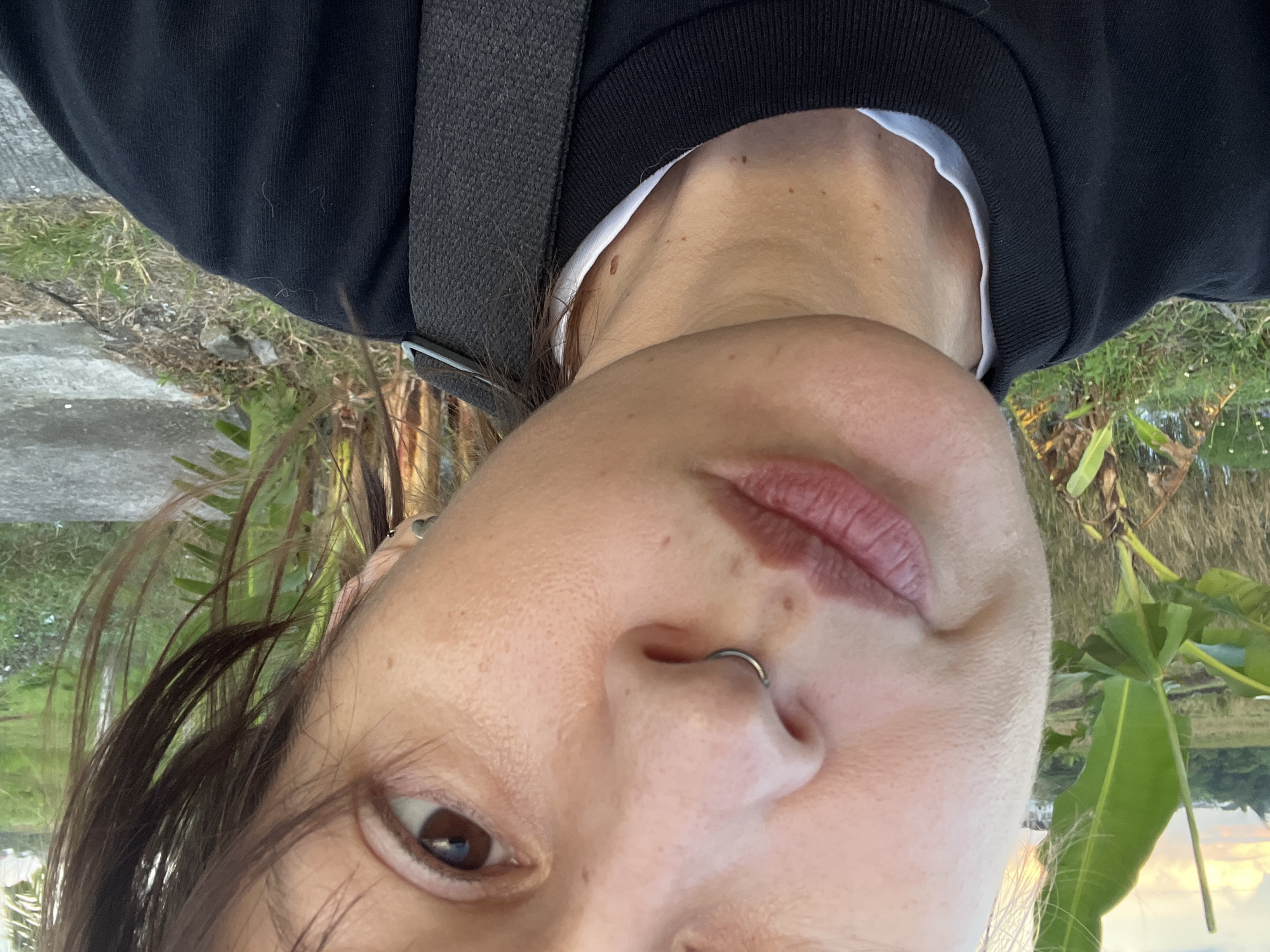 A close-up, upside-down portrait captures a person with brown hair and eyes looking directly at the camera. They wear a black long-sleeved top over a white undershirt, with a dark strap, possibly from a bag, visible across their shoulder. A silver septum piercing adorns their nose, and a small heart-shaped earring is visible in their right ear. Several moles or freckles are present on their neck and upper chest.
The background, which appears behind the person's head when the image is reoriented, depicts an outdoor scene in what appears to be North Kuta, Indonesia. Lush green tropical foliage and grass are prominent, with glimpses of a reflective surface, likely water, and a warm-toned sky, suggesting late afternoon or sunset. A grey concrete path or ground is also visible in the periphery. The overall impression is a casual outdoor selfie taken in a tropical environment. There is no visible text.