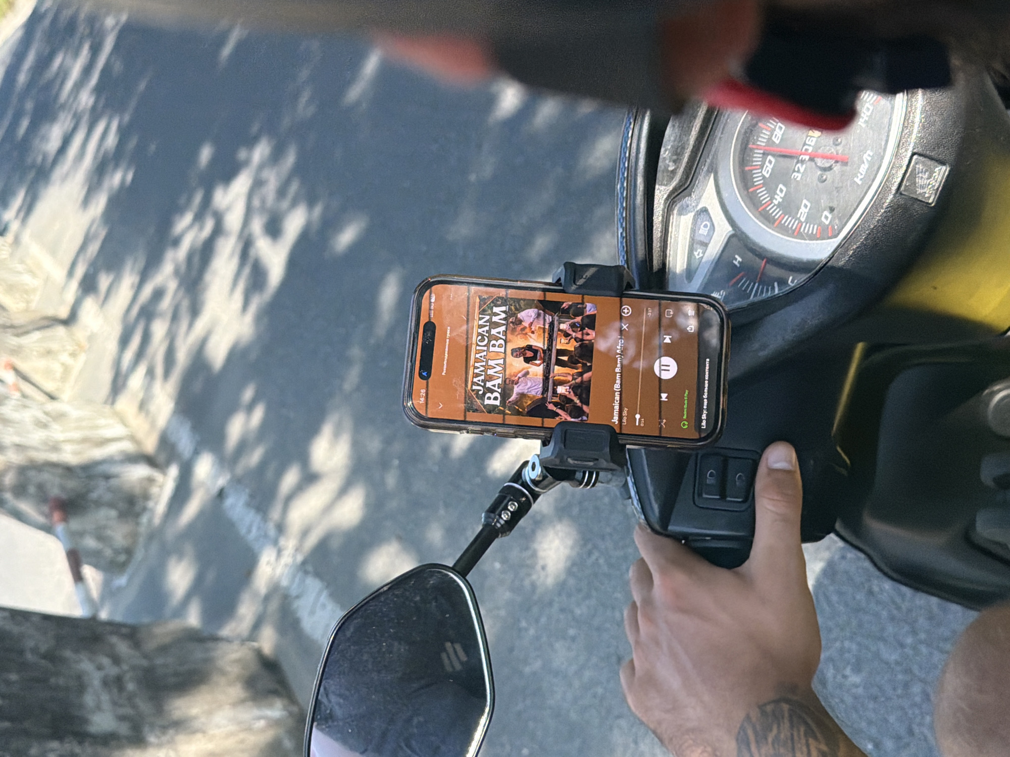 A first-person perspective shot from a motorcycle in Nam Nha Trang Ward, Vietnam, shows a hand gripping the handlebars. A smartphone mounted on the handlebars displays the album art and song information for "Jamaican Bam Bam" by Lilo Sky. The phone screen is illuminated, and the music player controls are visible. The road is paved, and the sun casts shadows from trees onto the asphalt. In the background, there are concrete structures and some vegetation. A motorcycle's speedometer, showing a speed of approximately 40 km/h, is also visible.