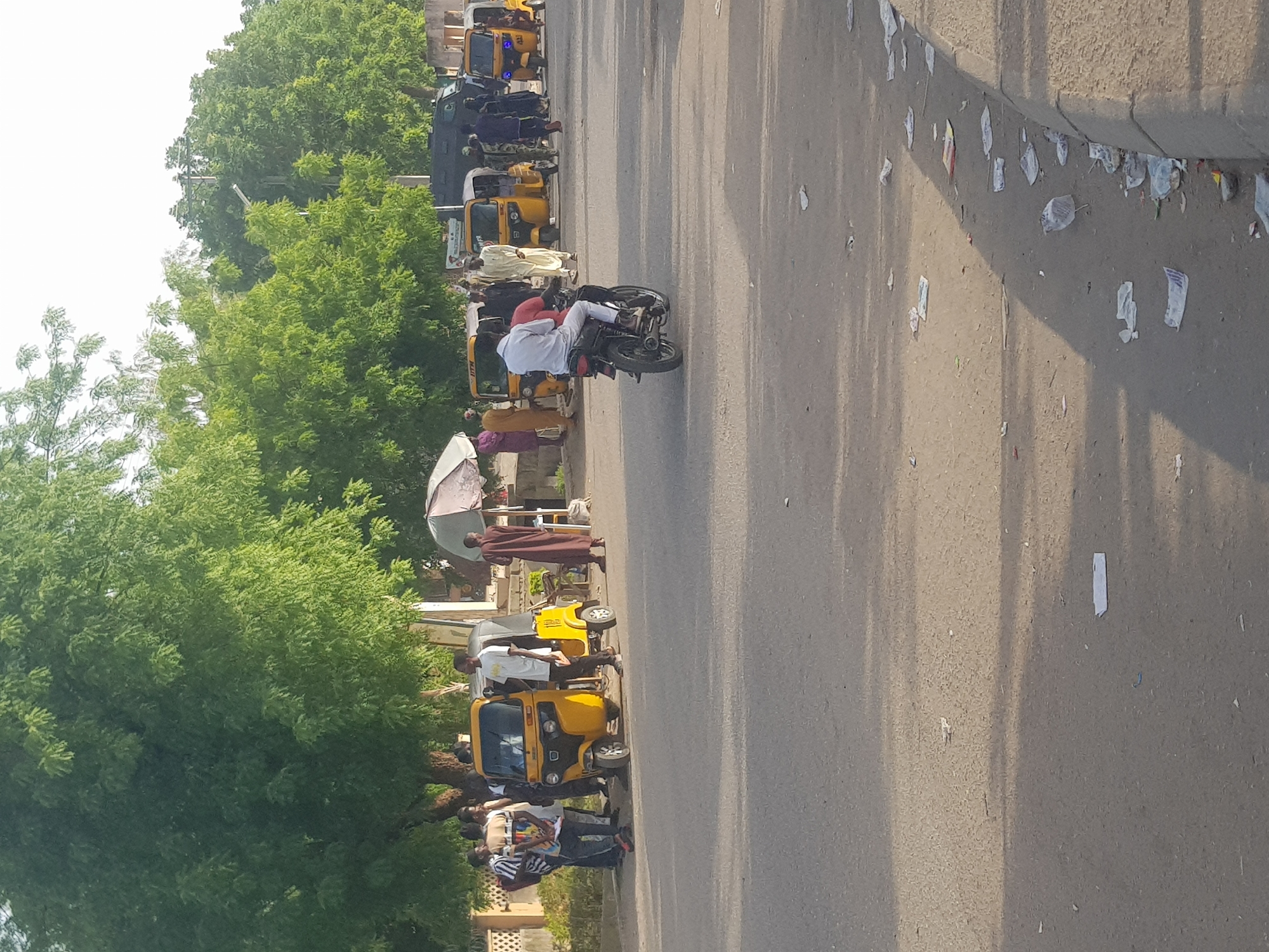 The image depicts a street scene in what appears to be Dabawa, Nigeria. A row of yellow auto-rickshaws (tuk-tuks) is parked along the left side of the street. People are standing around the tuk-tuks; some are dressed in traditional attire, while others wear casual clothing. One person rides a motorcycle. Trees line the left side of the street, with a building visible in the background. The street itself appears to be paved, with litter scattered along the curb.