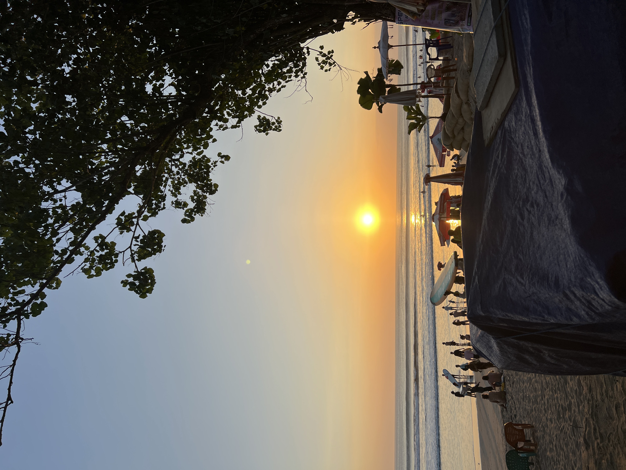 This image depicts a sunset scene on Kuta Beach, Bali, Indonesia. The sun, a bright orb, is setting over the ocean, casting a warm, golden glow across the sky and water. Gentle waves roll onto the sandy shore. A group of people are visible on the beach, some are standing near the water, while others are sitting or walking. Several surfboards are present, some are being carried by individuals, suggesting a surfing or surf lesson activity. In the foreground, a dark blue tarp covers what appears to be a table or stand, with chairs and sandbags positioned nearby. Beach umbrellas and palm trees add to the tropical atmosphere. The overall scene is serene, capturing the essence of a beach sunset with people enjoying the late afternoon light.