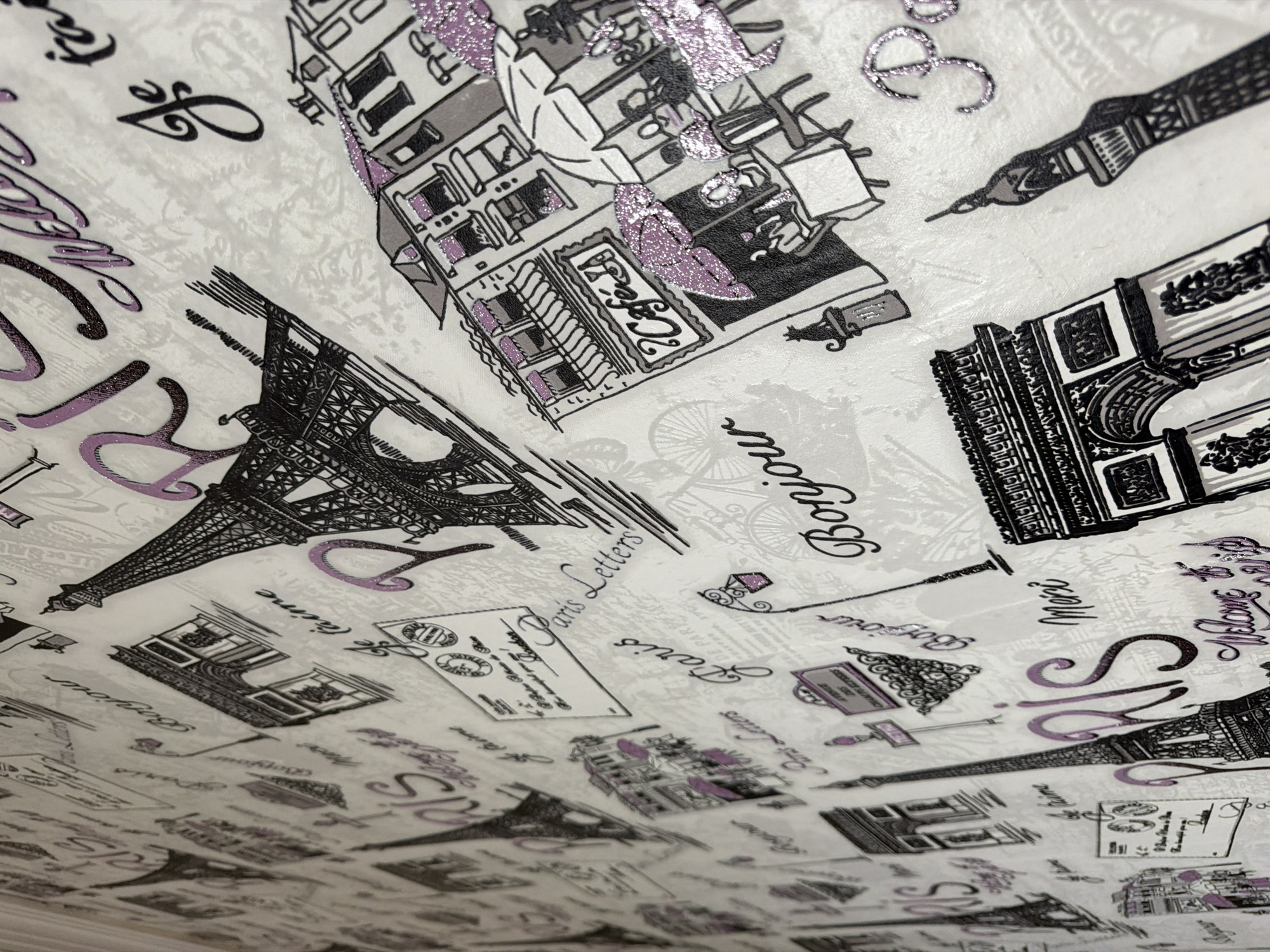 The image is a close-up, angled view of wallpaper with a Paris theme. The wallpaper is predominantly white with black and purple illustrations and text. The illustrations depict iconic Parisian landmarks such as the Eiffel Tower, the Arc de Triomphe, Parisian cafes with outdoor seating and umbrellas, and a street lamp. The text includes French phrases like "Paris," "Bonjour," "Merci," and "Je t'aime," as well as the word "Welcome." Some elements, particularly the purple accents on the umbrellas and text, appear to have a glittery or shimmering finish. The wallpaper has a textured appearance. There are no people or specific locations in Pazardzhik, Bulgaria visible in the image.
