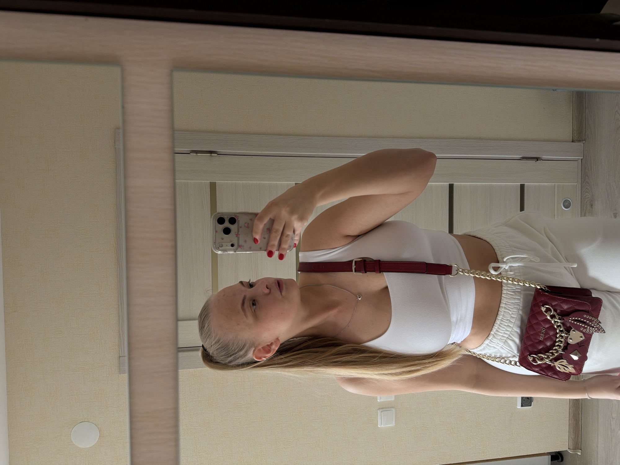 A young woman stands in front of a mirror, taking a selfie with her mobile phone. She wears a white crop top and white sweatpants, with a small, burgundy-colored purse slung across her body by a gold chain and burgundy strap. She has long, blonde hair pulled back in a low ponytail. The background consists of light-colored walls and a light-colored door with wood trim. The scene appears to be inside an apartment or house. The city of Izhevsk, Russia, is not visibly discernible in the image.
