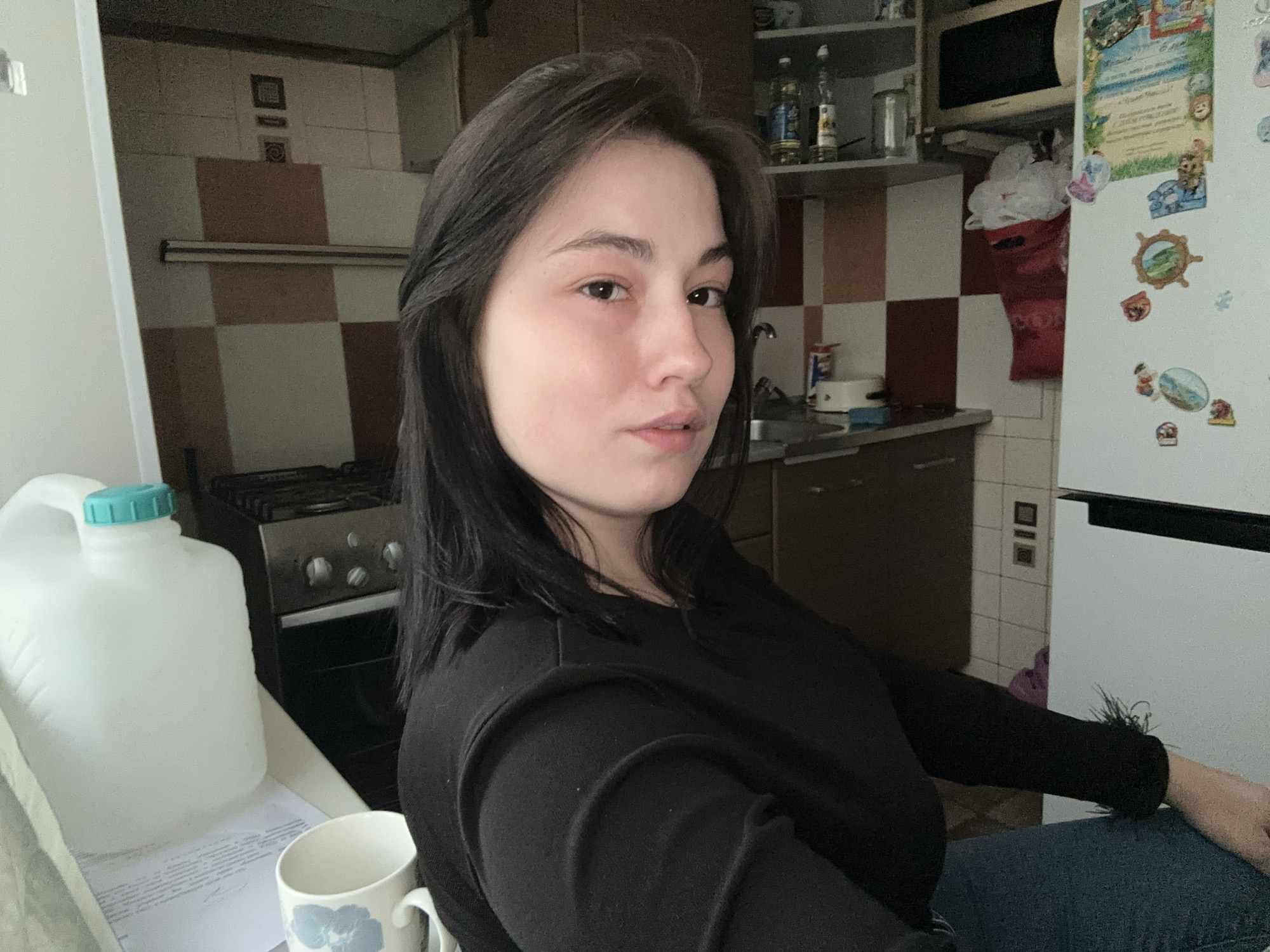 A young woman sits in a kitchen with checkered tile walls, likely in an apartment. The kitchen has a stove, a refrigerator with magnets, and cabinets with a microwave on top. A white pitcher with a turquoise lid sits on a counter next to a white mug with a blue flower design. The woman is wearing a black long-sleeved shirt and blue jeans. The location is presumed to be Chaikovsky, Russia.