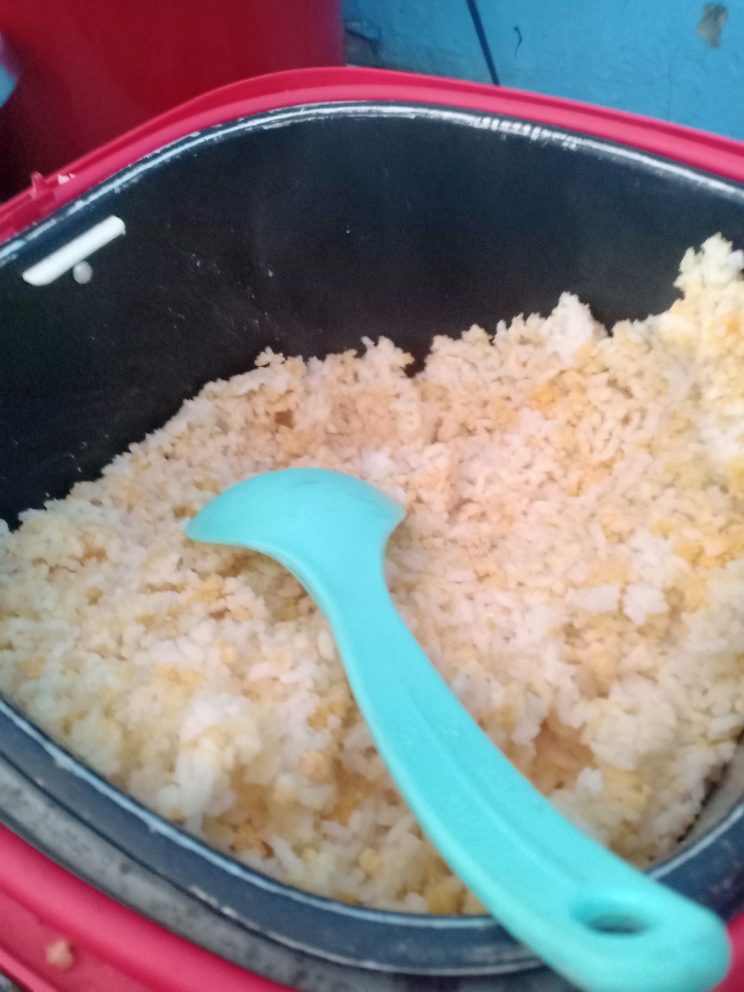 A close-up, high-angle shot shows a turquoise plastic spoon resting in a pot of white rice with some yellow bits mixed in. The rice is in a black inner pot of a rice cooker, which is set inside a red outer casing. The background is blurry, with hints of a red object on the left and a blue surface on the top right. The scene appears to be in a kitchen or dining area in Sumberrejo, Indonesia.