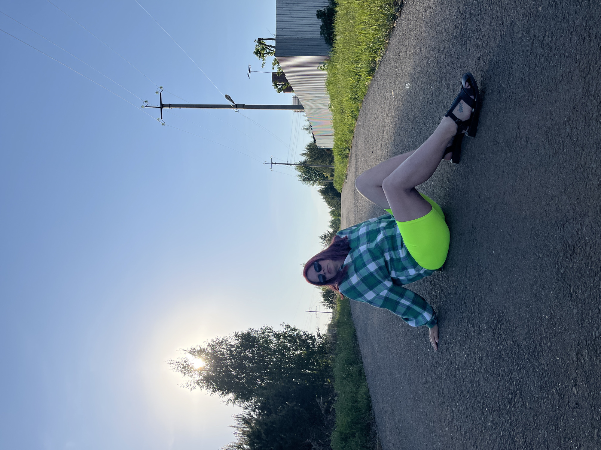 A woman is lying on a paved road in Chaikovsky, Russia. She is wearing sunglasses, a green and white plaid shirt, and bright green shorts. She has dark sandals on her feet. A power line runs along the road and there are trees and shrubs nearby. The sun is shining brightly in the background.