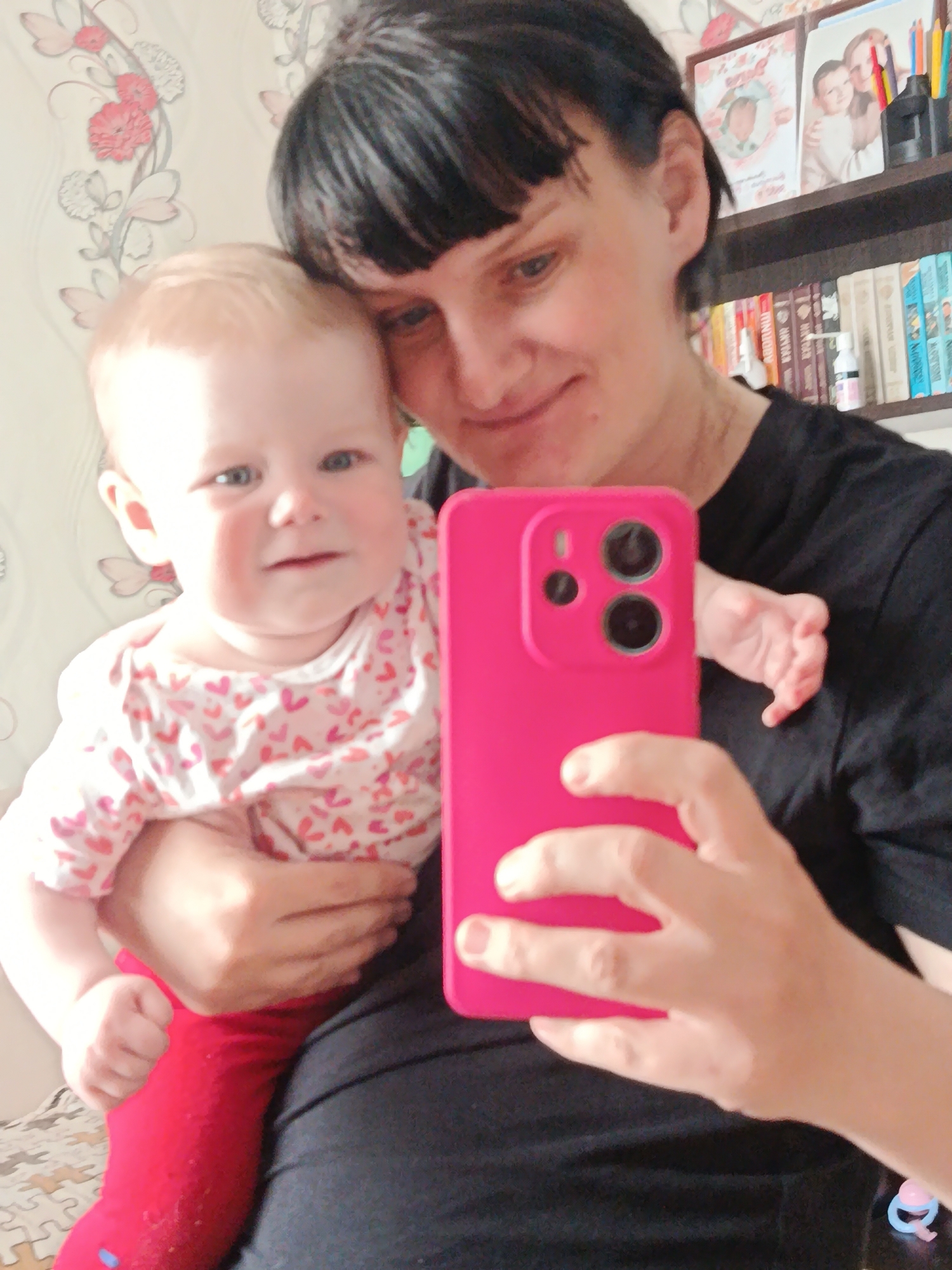 A woman holds a baby in her arms while taking a selfie with a pink smartphone. The woman has short, dark hair and is wearing a black shirt. The baby has light hair and is wearing a white shirt with small red hearts and red pants. The background includes floral wallpaper and a bookshelf. The location is Unknown, Unknown.