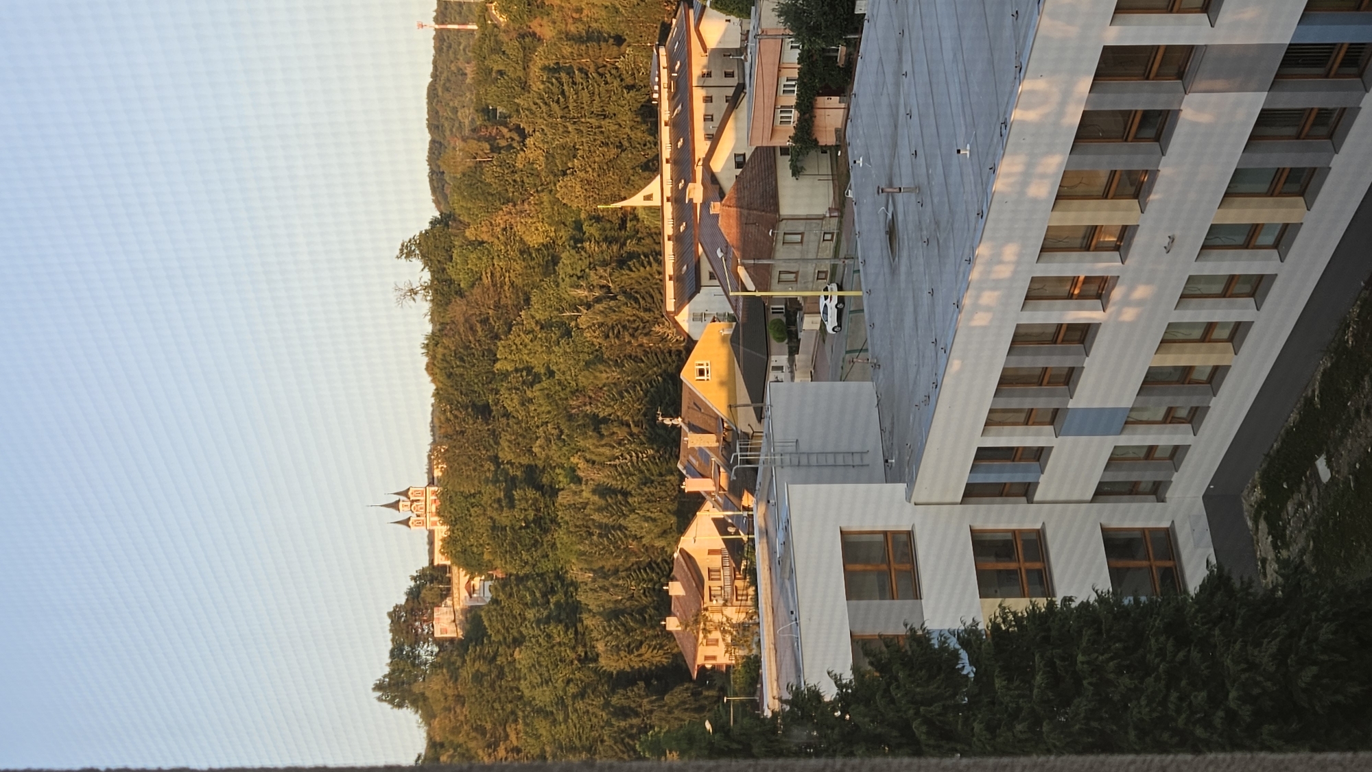 The image shows a view of a residential area with a church on a hill in the background. The church has a distinctive red and white facade and two spires. The foreground is dominated by a modern white building with large windows. The buildings in the middle ground are typical houses with tiled roofs. The scene is set in a town with a lot of greenery, indicating it might be a suburban or semi-rural area. The lighting suggests it is either early morning or late afternoon, with the sun casting long shadows.
The presence of the church with its unique architecture, and the overall urban landscape with its blend of older and modern buildings, strongly suggests the location is the vicinity of the Holy Trinity Church in Prešov, Slovakia. The church is a prominent landmark on a hill overlooking the city. The modern building in the foreground could be a school or administrative building, common in city centers or developed residential areas. The houses in the middle ground are characteristic of European residential architecture.