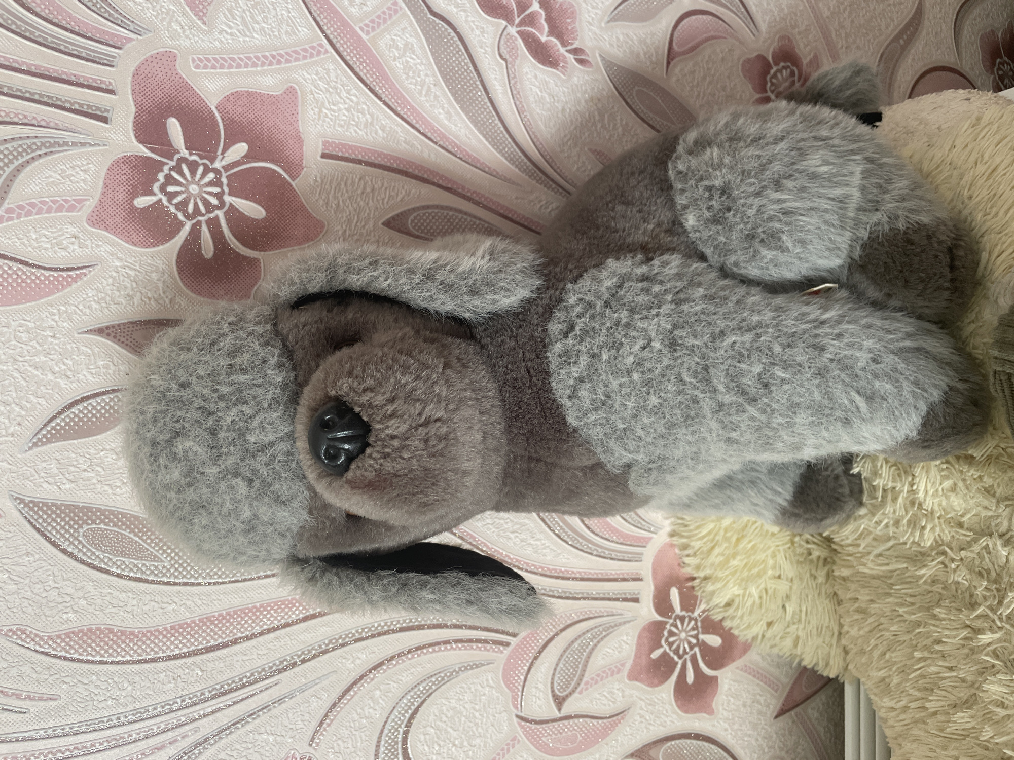 A plush toy dog, appearing to be a gray poodle, sits on a beige fluffy surface in front of a pink and white floral patterned wallpaper. The dog has floppy ears and a darker gray chest. The wallpaper has a textured finish and features repeating floral motifs and leaf-like shapes. The lighting is soft and natural. The scene is indoors, with no visible indications of a specific city or outdoor environment.