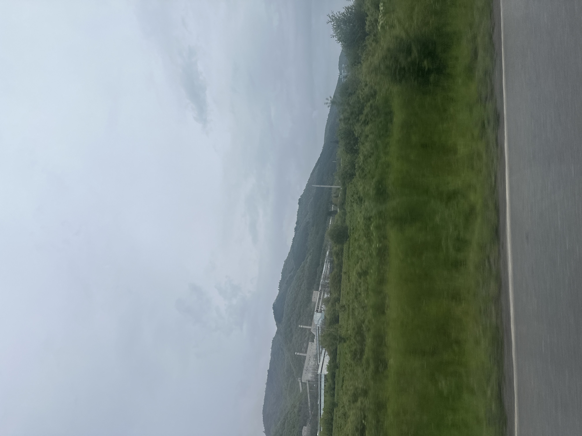 The image shows a landscape view in Mirkovo, Bulgaria, taken from a moving vehicle. A mountain range is visible in the background, covered with dense green vegetation. In the midground, there is a large industrial complex with tall structures and smokestacks. The sky is overcast and gray, suggesting a cloudy or rainy day. The foreground shows the edge of a road or highway.