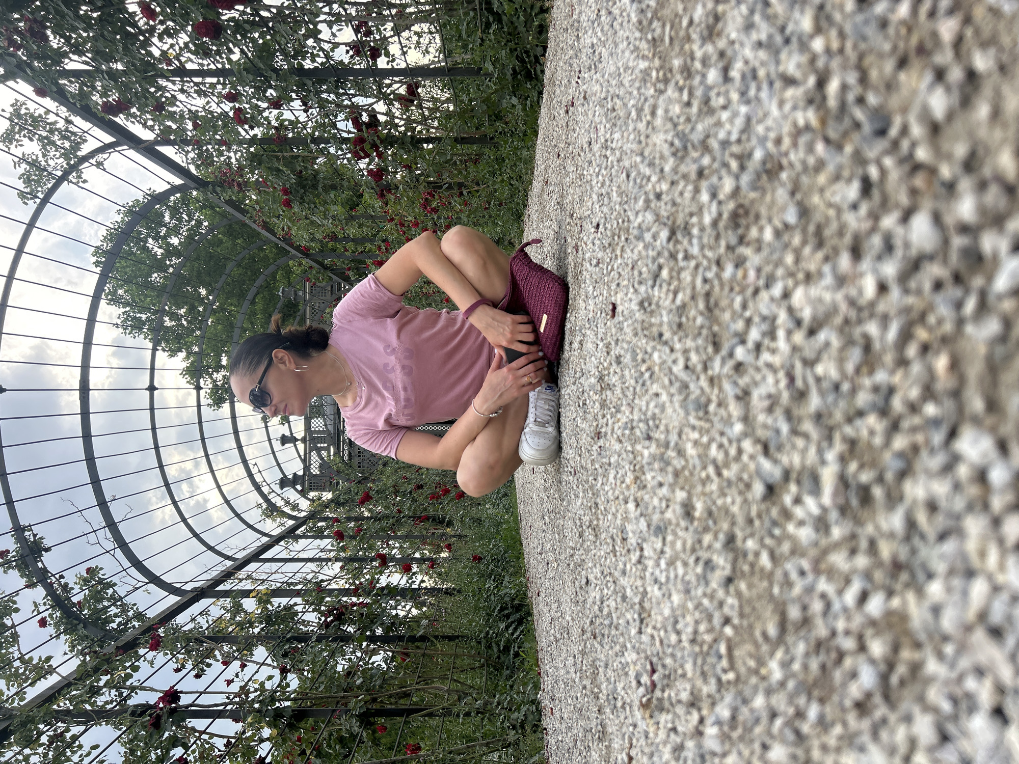 A woman is crouched on a gravel path beneath a rose-covered trellis. She wears a pink t-shirt, white sneakers, and sunglasses. A maroon clutch rests on the gravel beside her. The trellis, made of dark metal, arches over the path and is draped with green foliage and red roses. Beyond the trellis, trees and a cloudy sky are visible. The scene appears to be outdoors, possibly in a park or garden, with the gravel path suggesting a manicured outdoor space. The lighting suggests it might be daytime, possibly late afternoon or early evening, given the soft light. The woman's posture and relaxed demeanor suggest she is taking a break or enjoying the surroundings. The overall impression is one of a peaceful moment in a botanical setting.